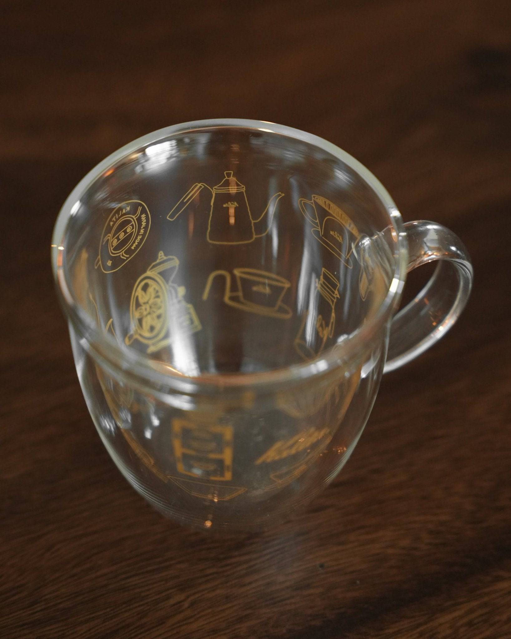 Kalita double-walled glass mug with coffee-themed illustrations, featuring a kettle, grinder, and filter, on a wooden surface. Clear glass showcases intricate gold designs.