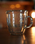 Kalita double-walled glass mug with coffee equipment illustrations etched on the surface, placed on a wooden surface with warm background lighting