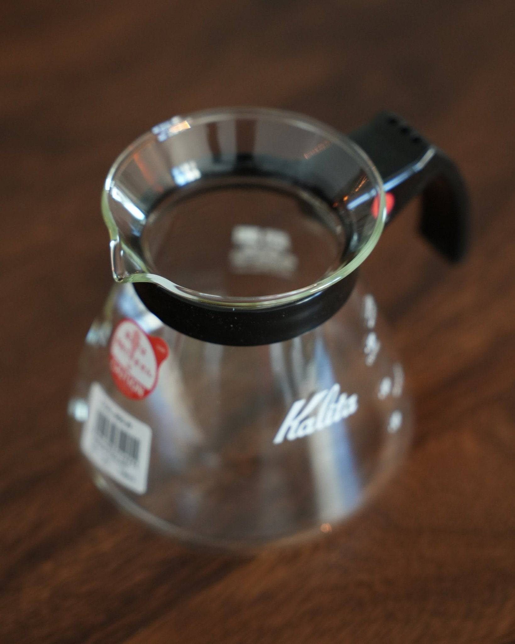 Kalita 500 Server N glass coffee pot with heat-resistant body, black handle, and wide mouth, sitting on a wooden surface. Clear glass shows Kalita logo. Red tag visible, indicating brand authenticity and quality.