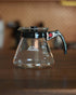 Kalita 500 Server N glass coffee carafe with black lid and handle, placed on wooden surface. Clear glass body shows Kalita logo and measurement markings. Ideal for brewing and serving coffee or tea.