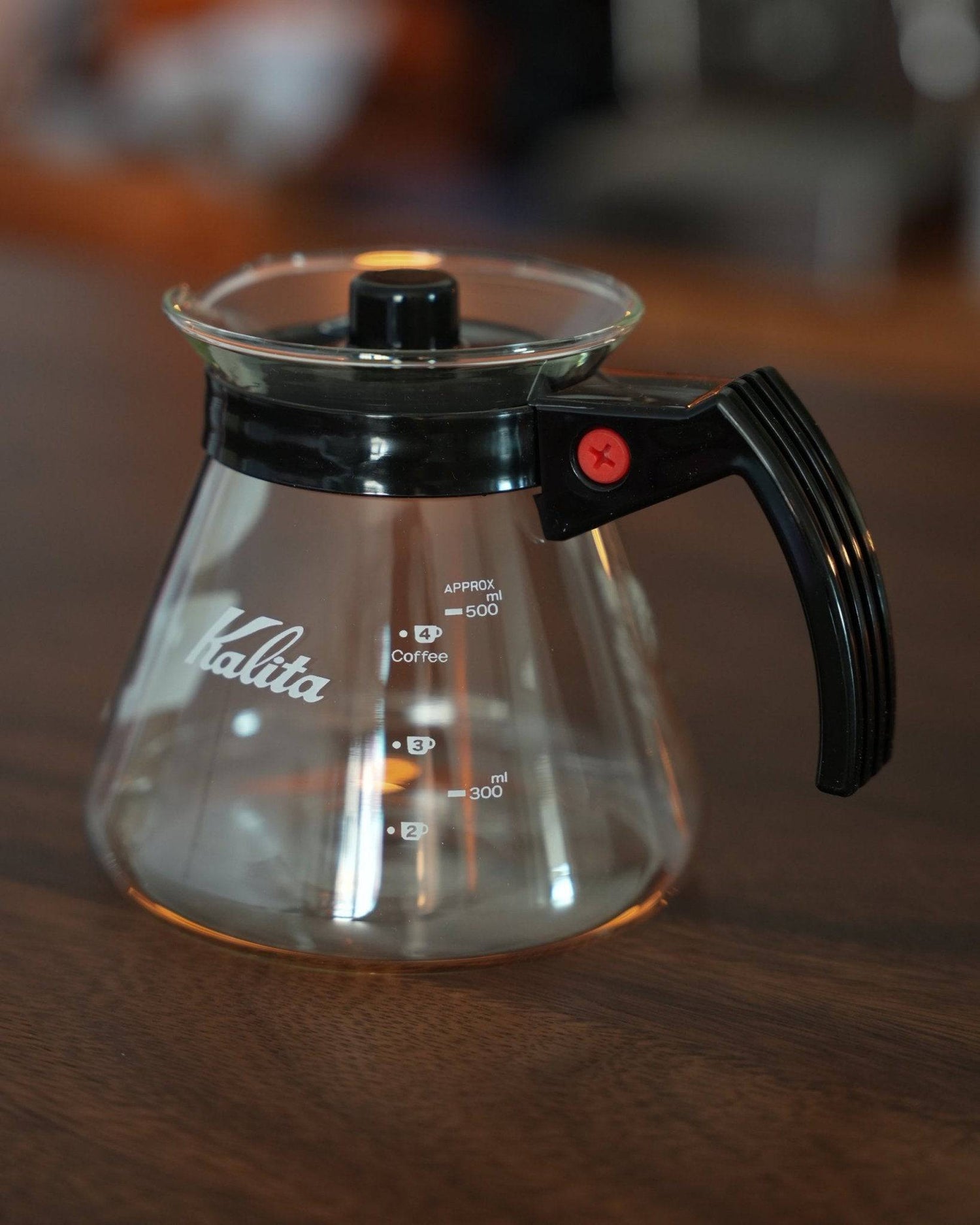 Kalita 500 Server N heat-resistant glass coffee server, 500ml capacity, with black handle and lid, measurement markings, on wooden surface, warm lighting