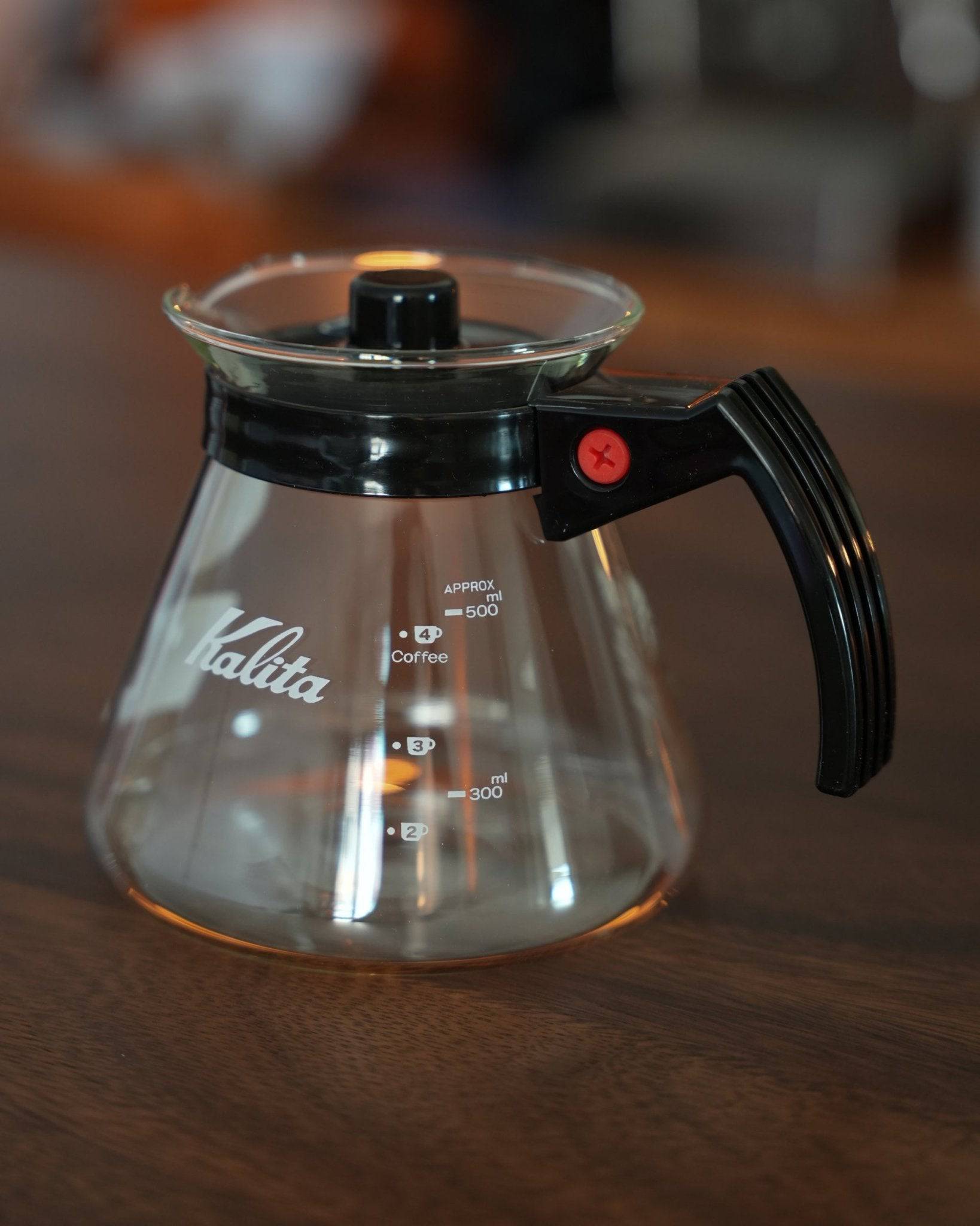 Kalita 500 Server N heat-resistant glass coffee server, 500ml capacity, with black handle and lid, measurement markings, on wooden surface, warm lighting