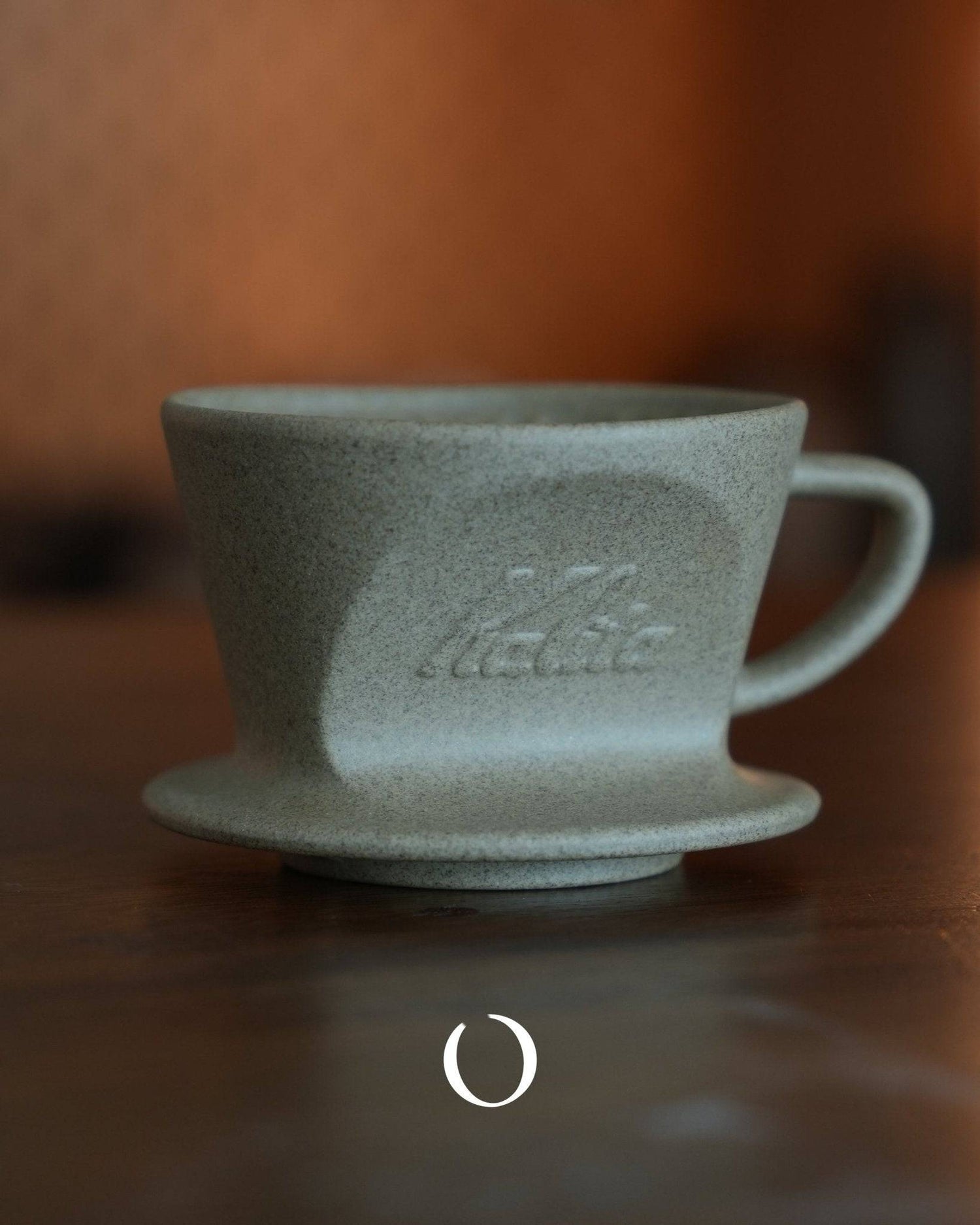 Kalita SG-101/SG-102 sandstone clay Hasami-yaki coffee dripper, gray-green textured surface with embossed Kalita logo, conical shape with handle, sitting on wooden surface against warm brown background
