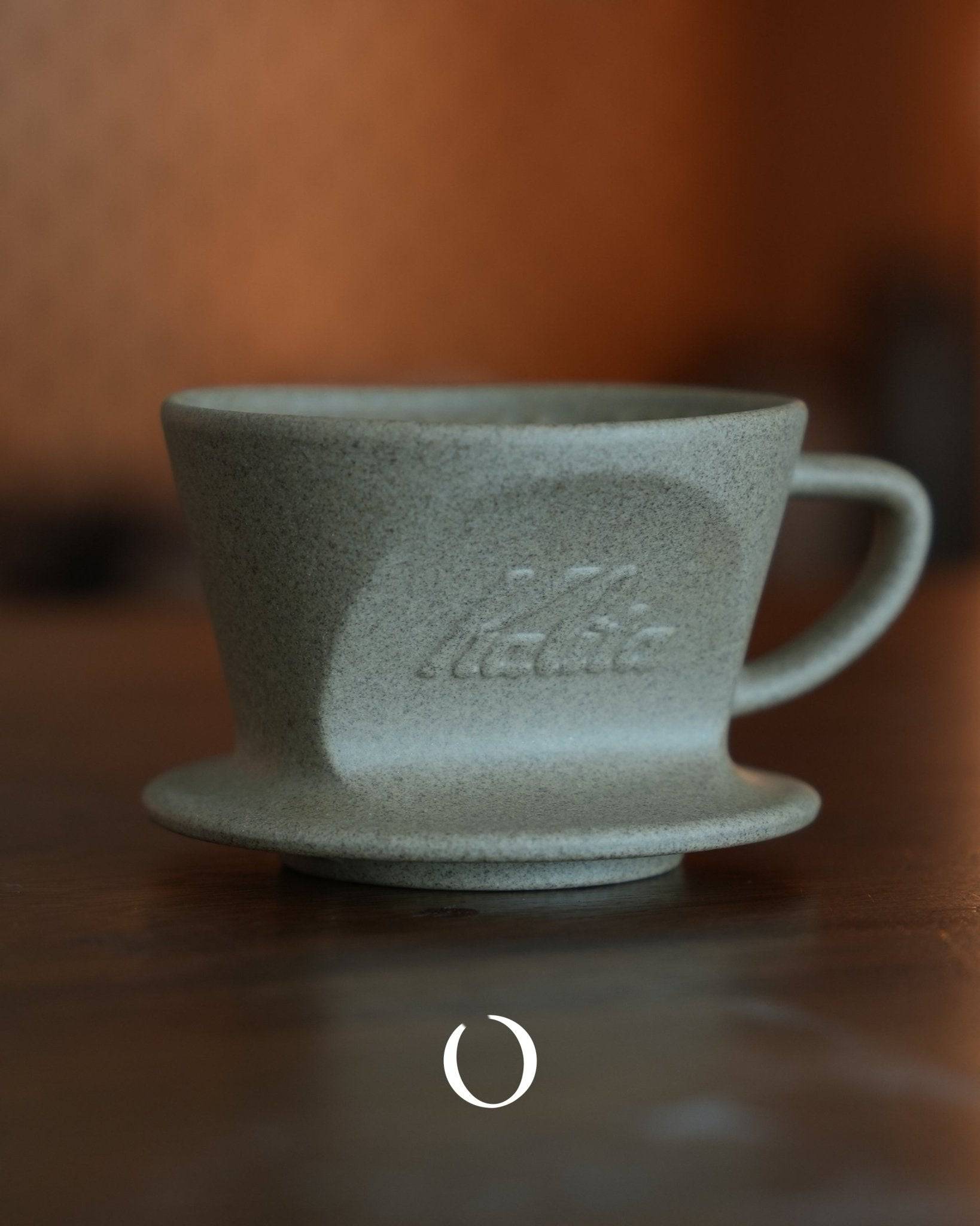 Kalita SG-101/SG-102 sandstone clay Hasami-yaki coffee dripper, gray-green textured surface with embossed Kalita logo, conical shape with handle, sitting on wooden surface against warm brown background