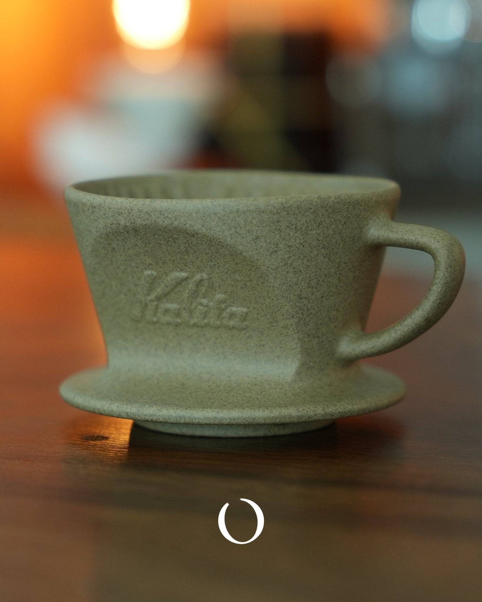 Kalita sandstone ceramic pour-over coffee dripper, sage green color, textured surface, with Kalita logo, on wooden surface with warm lighting, showcasing Japanese craftsmanship and design for optimal coffee brewing