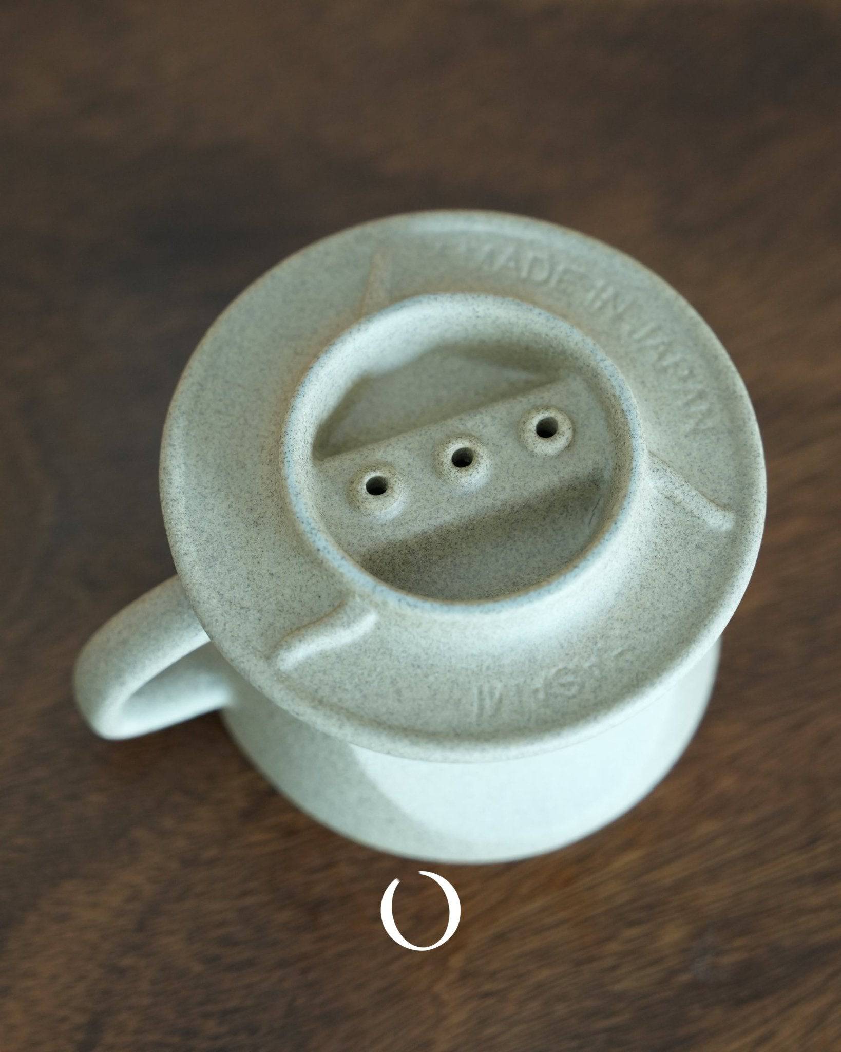 Kalita SG series sandstone clay Hasami-yaki coffee dripper, top view showing three filter holes, made in Japan text visible, light gray ceramic texture, conical shape with handle, on wooden surface