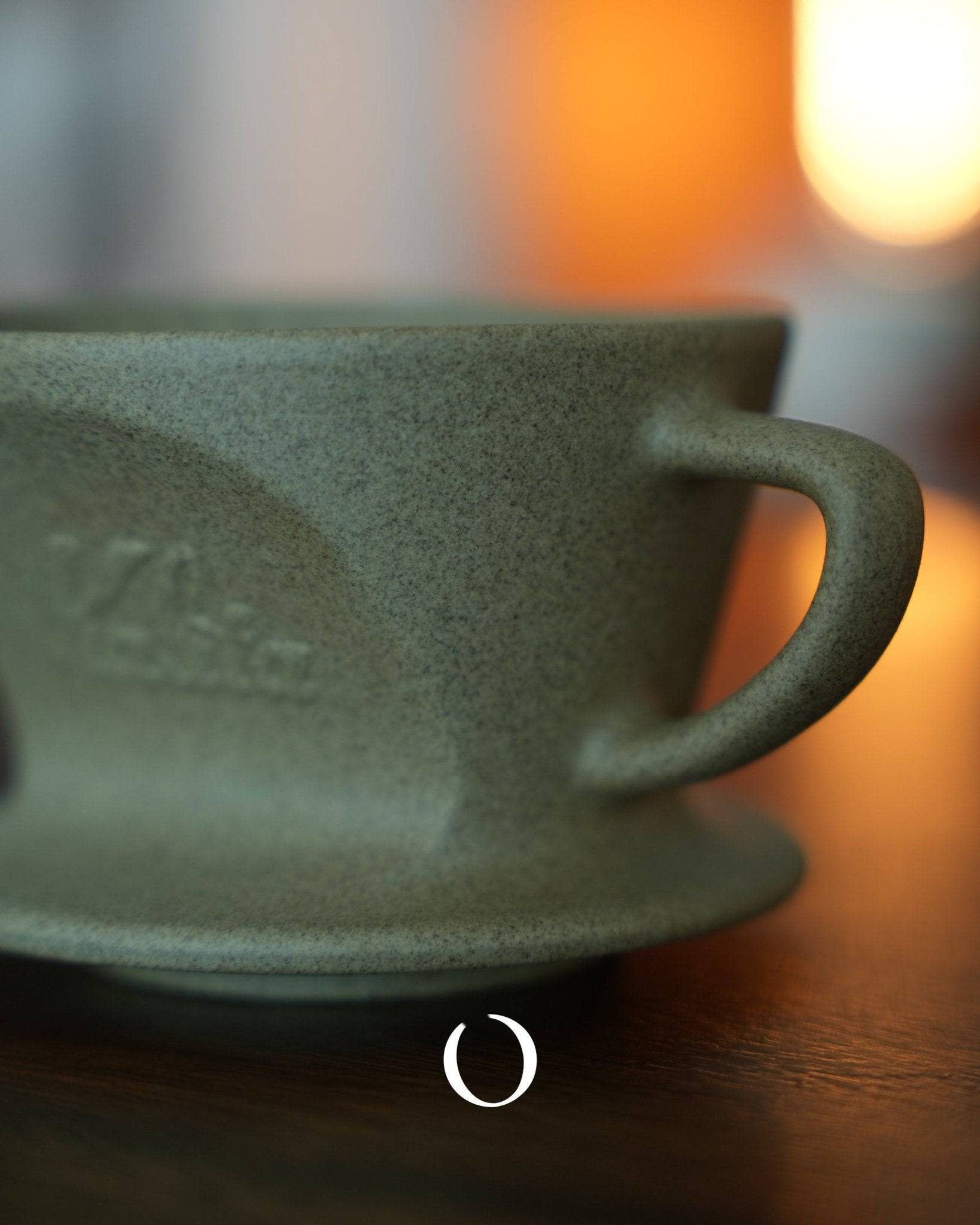 Close-up of Kalita SG series sandstone clay coffee dripper, showcasing its textured surface and elegant handle. Warm lighting in background highlights the dripper&