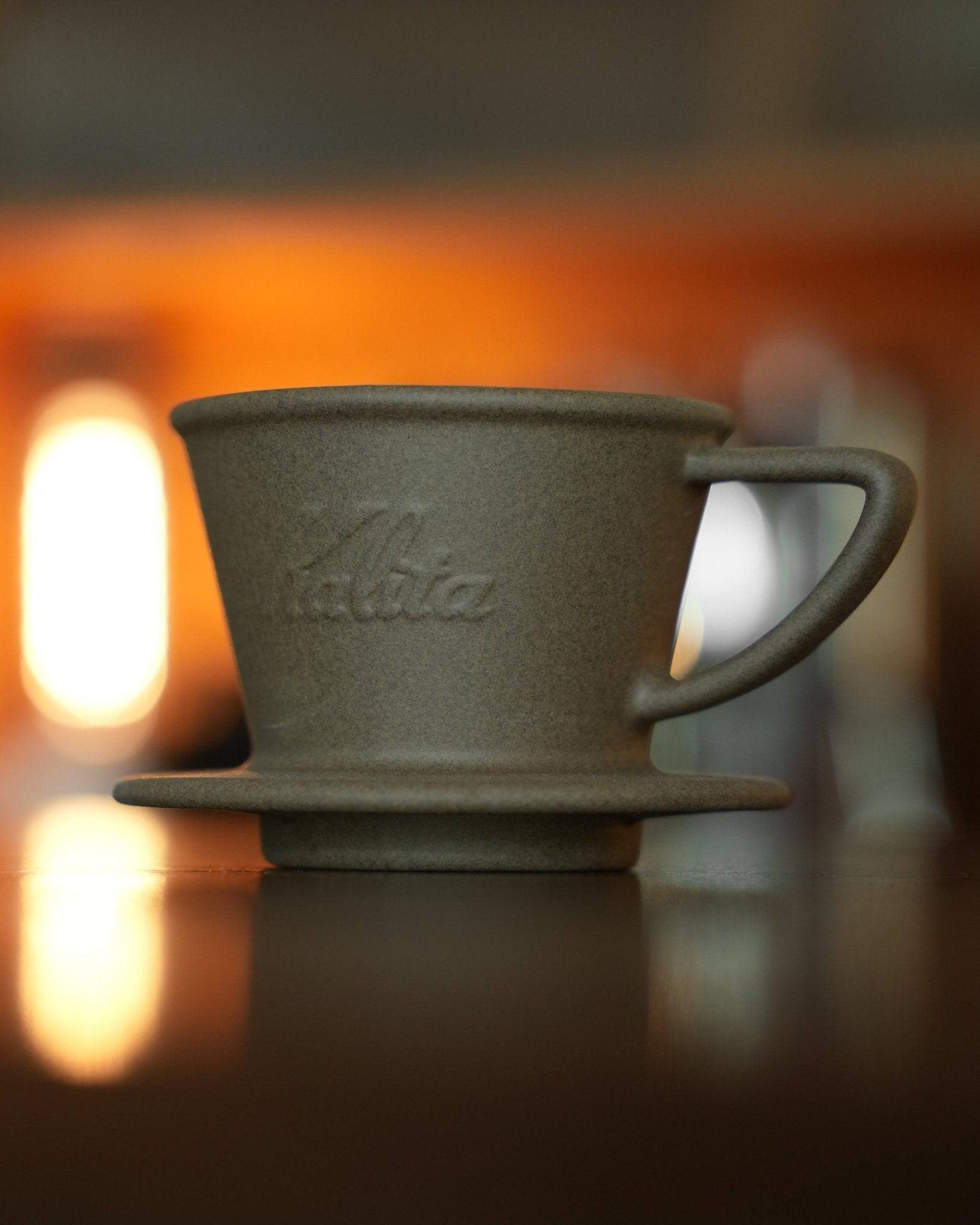 Kalita SG-155 Hasami-yaki sandstone coffee dripper, matte gray ceramic pour-over cone with handle, traditional Japanese craftsmanship, on reflective surface with warm background lighting