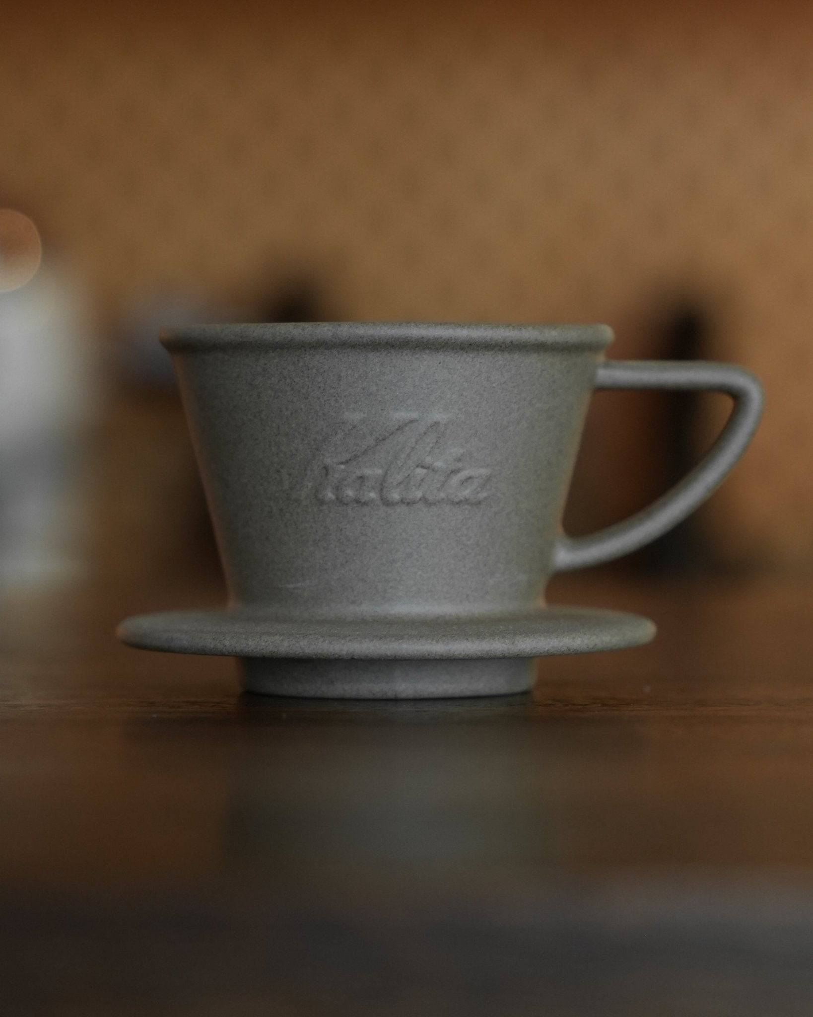 Kalita SG-155 Hasami pottery coffee dripper on wooden surface, matte gray ceramic with embossed logo, conical shape with handle, traditional Japanese craftsmanship meets modern coffee brewing technology