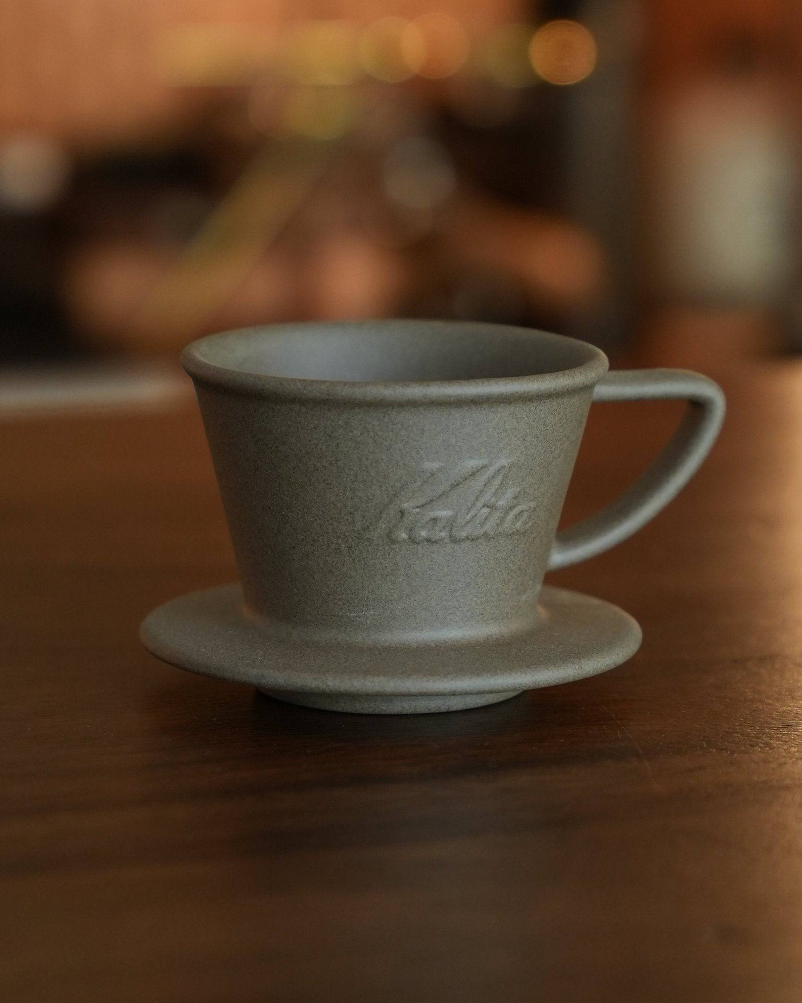 Kalita SG-155 Hasami-yaki sandstone coffee dripper, grey ceramic pour-over cup with saucer, rustic texture, embossed Kalita logo, on wooden surface with blurred background