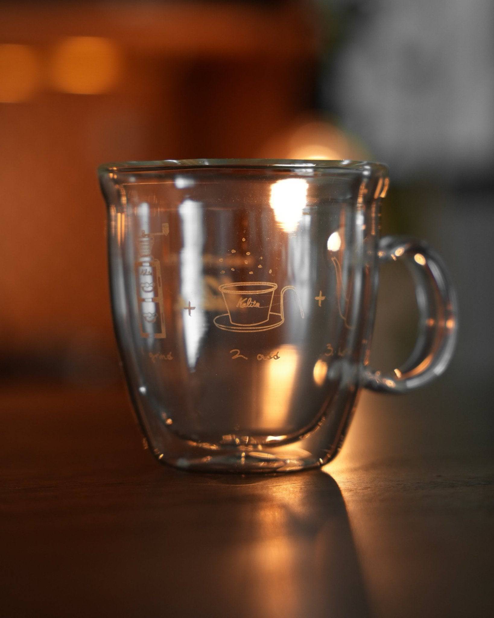 Kalita double-walled glass mug with etched coffee cup design, illuminated by warm ambient light, showcasing transparency and craftsmanship