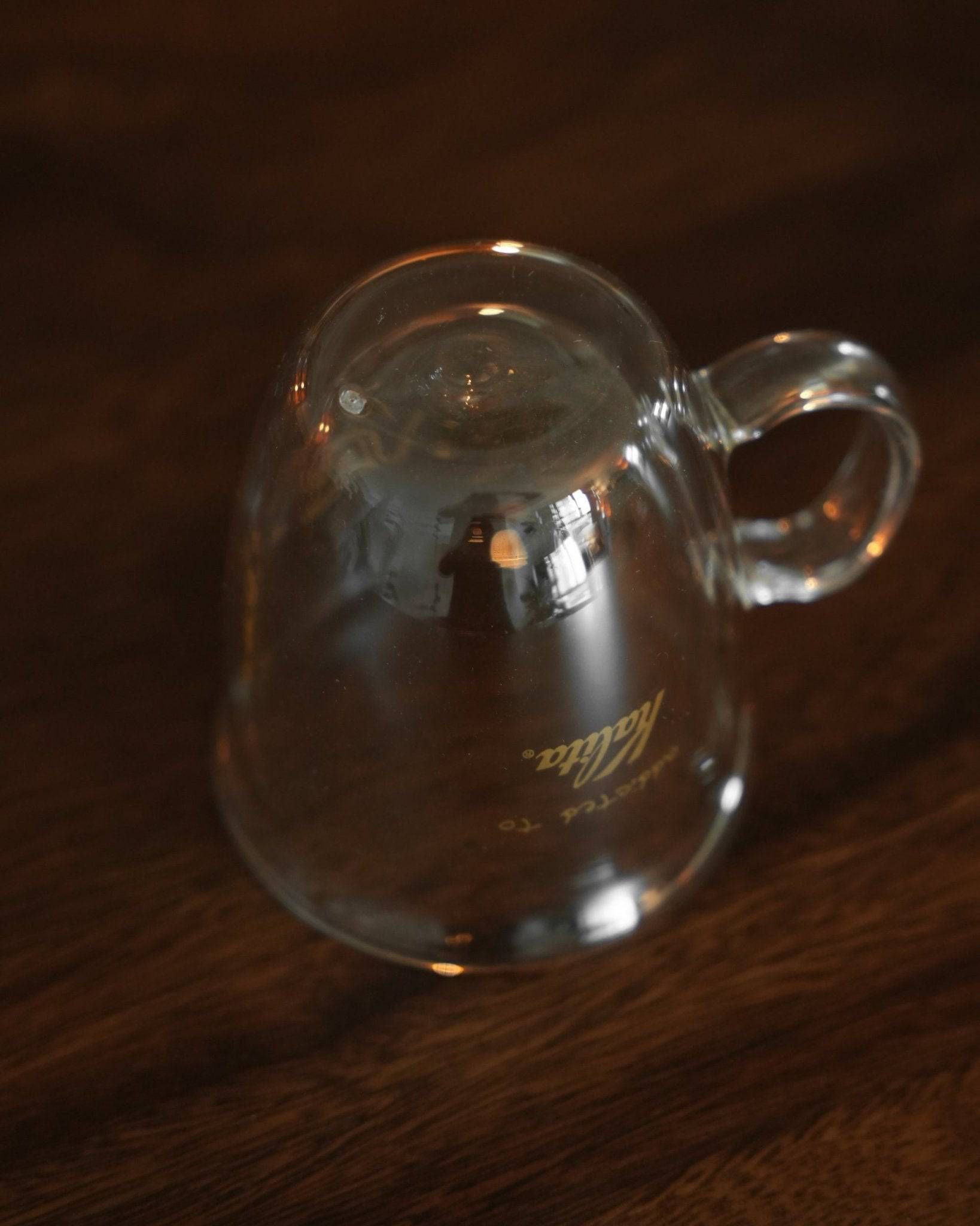 Kalita double-walled glass mug on wooden surface, showcasing transparent design with gold logo, curved handle, and light reflections highlighting its sleek form