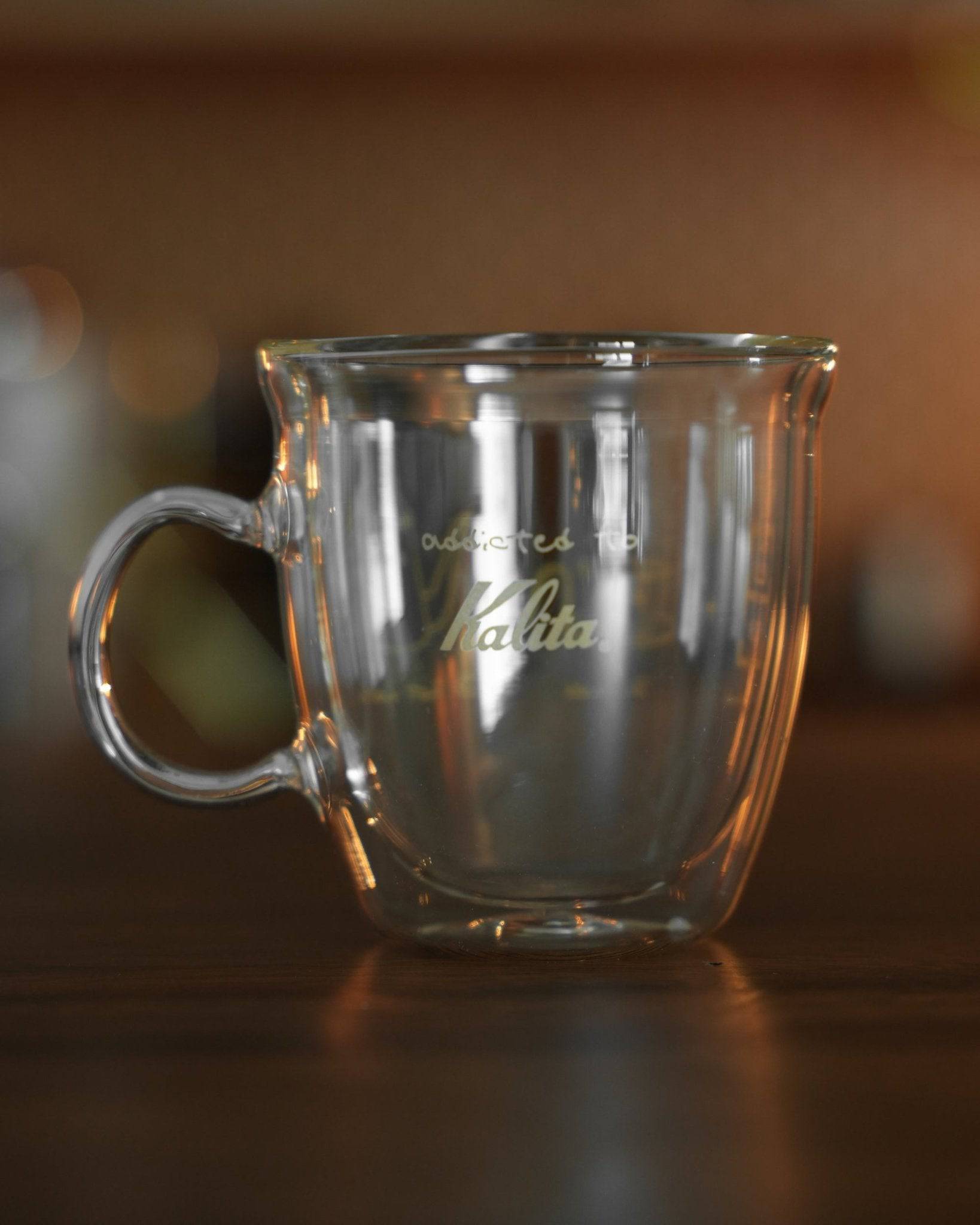 Kalita double-walled glass mug with logo, transparent design showcasing insulation, curved handle, sitting on wooden surface with soft background lighting