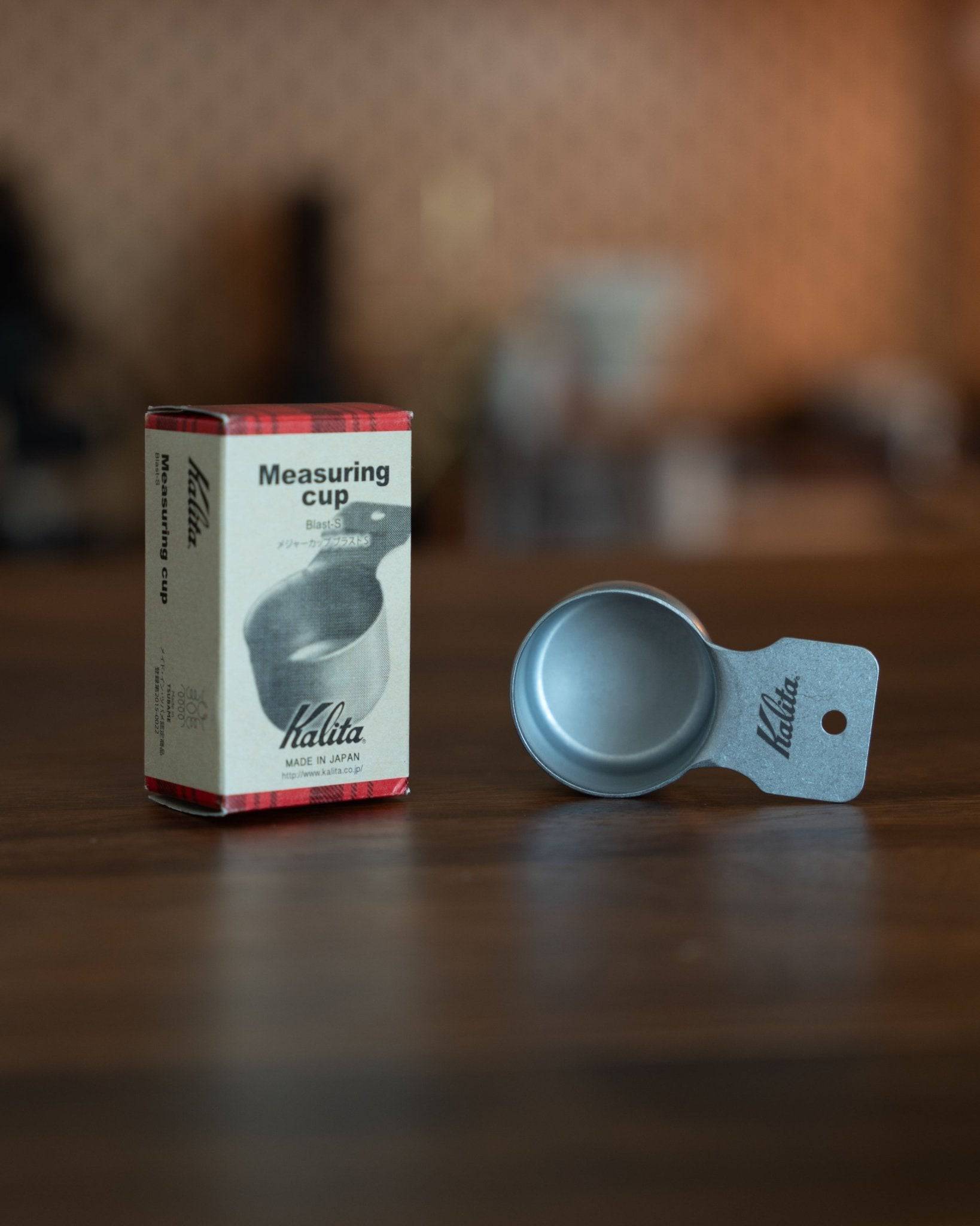 Kalita stainless steel coffee measuring scoop 10g (S Size) next to its packaging box on a wooden surface, showcasing the product&