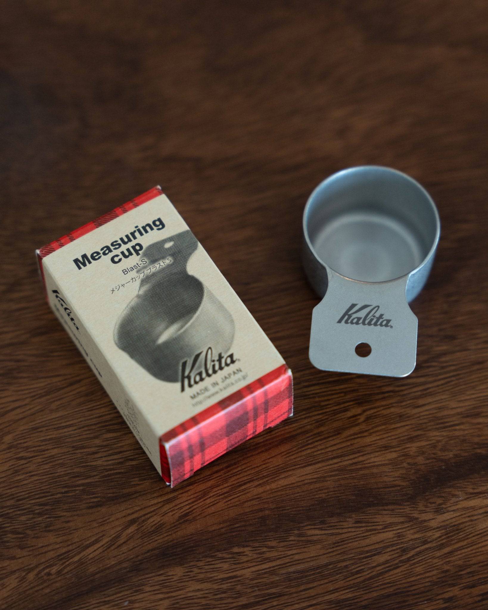 Kalita stainless steel coffee measuring cup and its packaging box on wooden surface, showcasing precision coffee tools for brewing enthusiasts