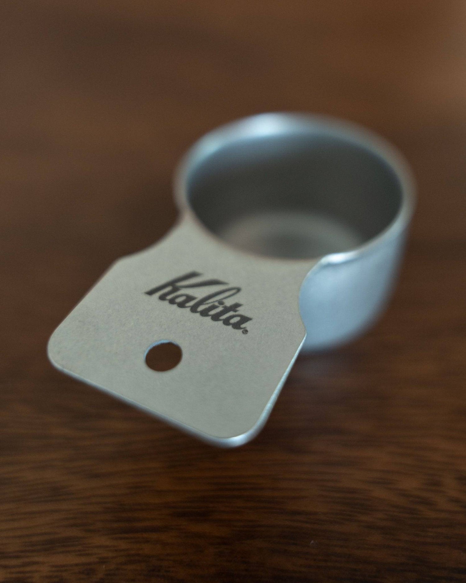 Kalita stainless steel coffee measuring spoon 10g (S Size) on wooden surface, showcasing its sleek design, matte finish, and branded handle with a small hole