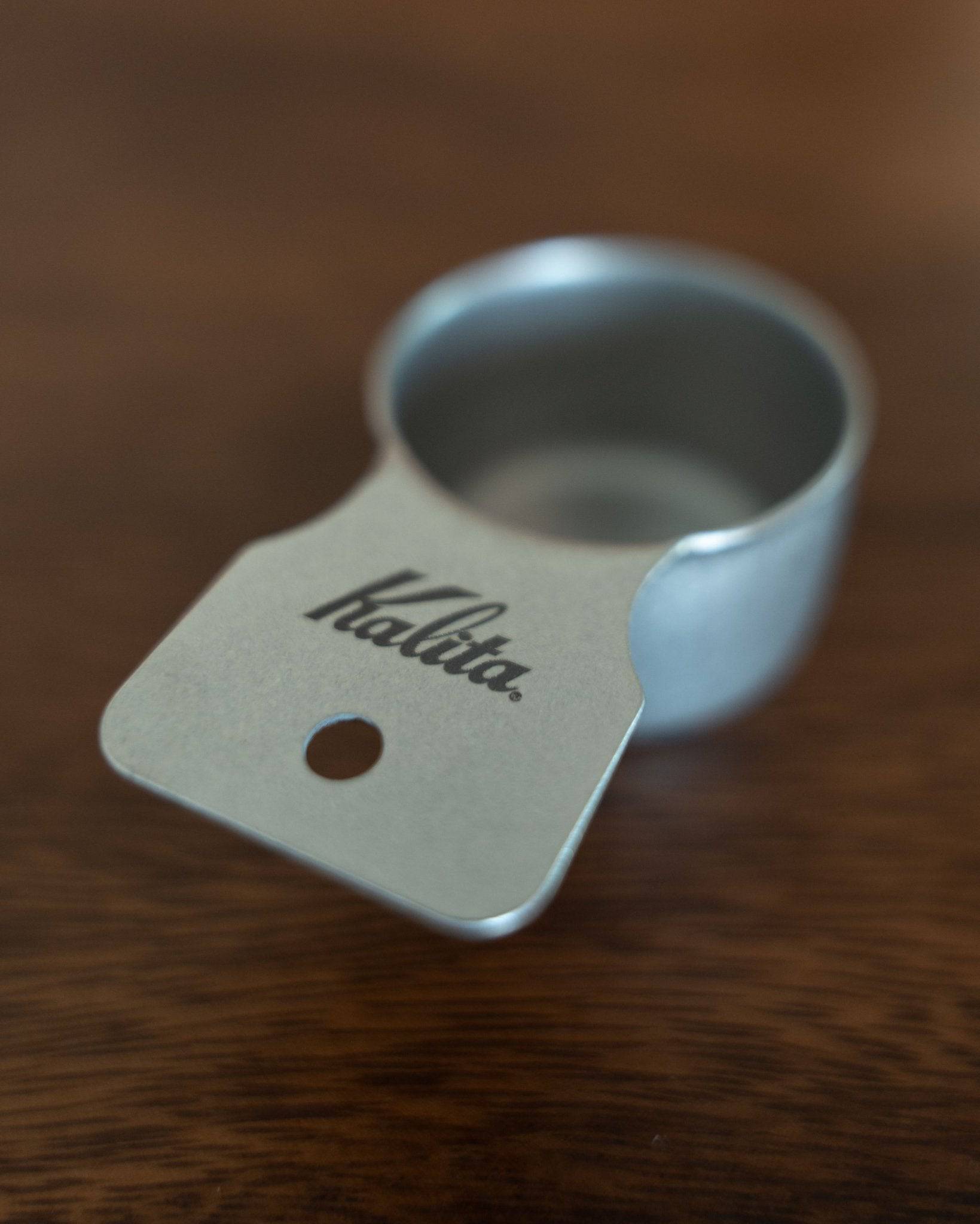 Kalita stainless steel coffee measuring spoon 10g (S Size) on wooden surface, showcasing its sleek design, matte finish, and branded handle with a small hole