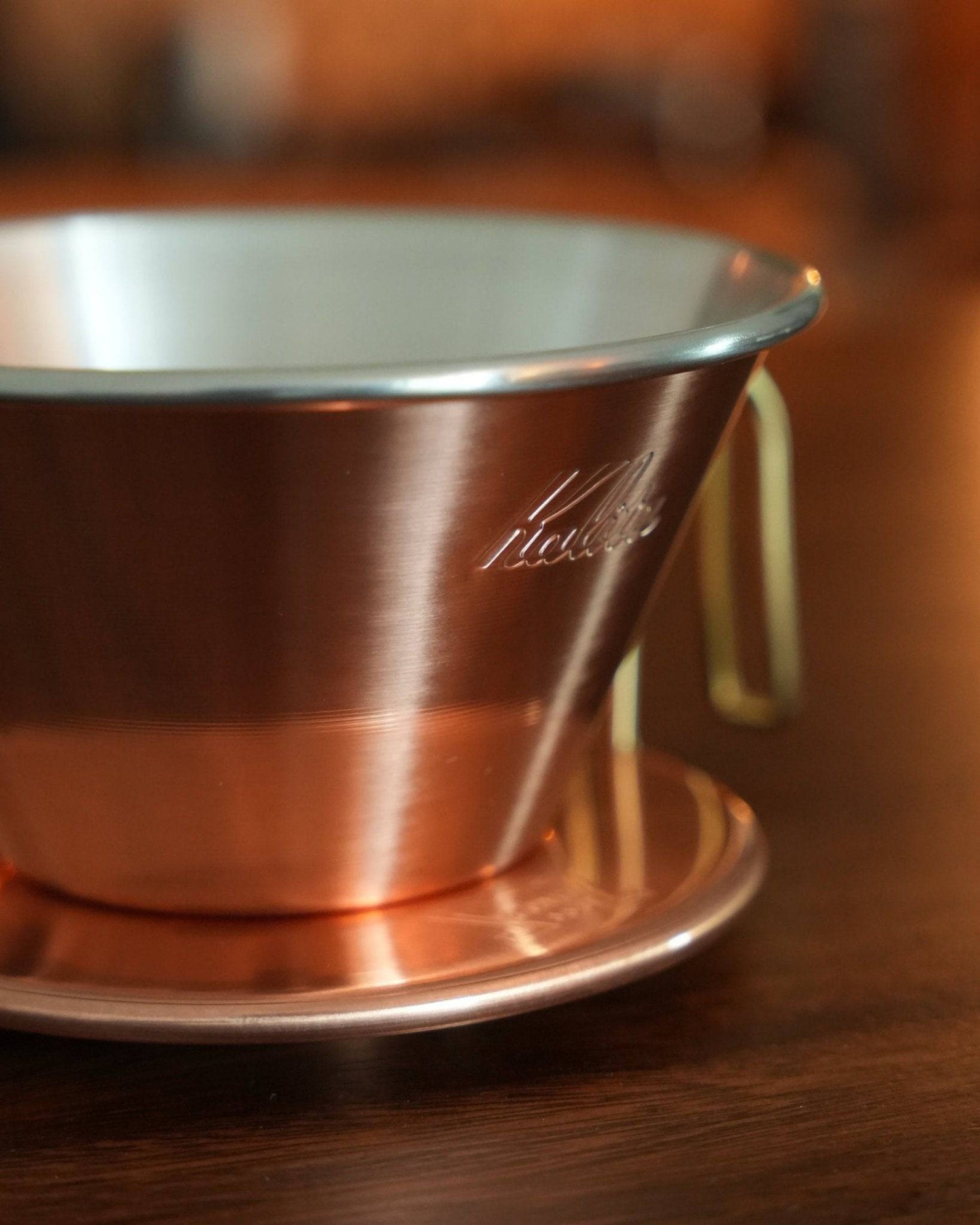 Kalita & TSUBAME copper coffee dripper with elegant design, showcasing its shiny copper exterior, silver rim, and Kalita branding engraved on the side, positioned on a wooden surface with warm lighting highlighting its premium craftsmanship