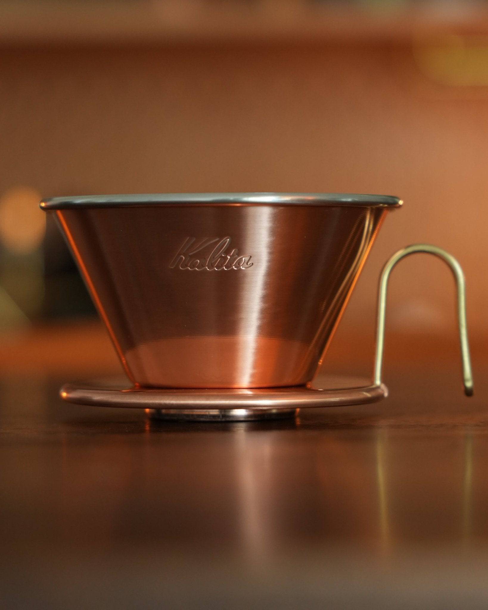 Copper Kalita pour-over coffee dripper with three-hole design on wooden surface, showcasing its elegant conical shape and high-quality craftsmanship for precise coffee brewing