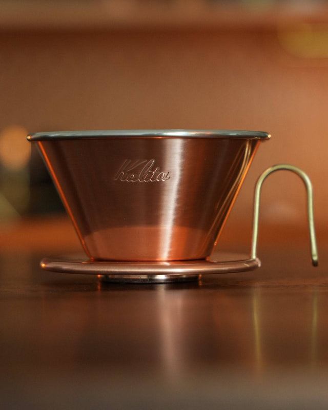 Copper Kalita pour-over coffee dripper with three-hole design on wooden surface, showcasing its elegant conical shape and high-quality craftsmanship for precise coffee brewing