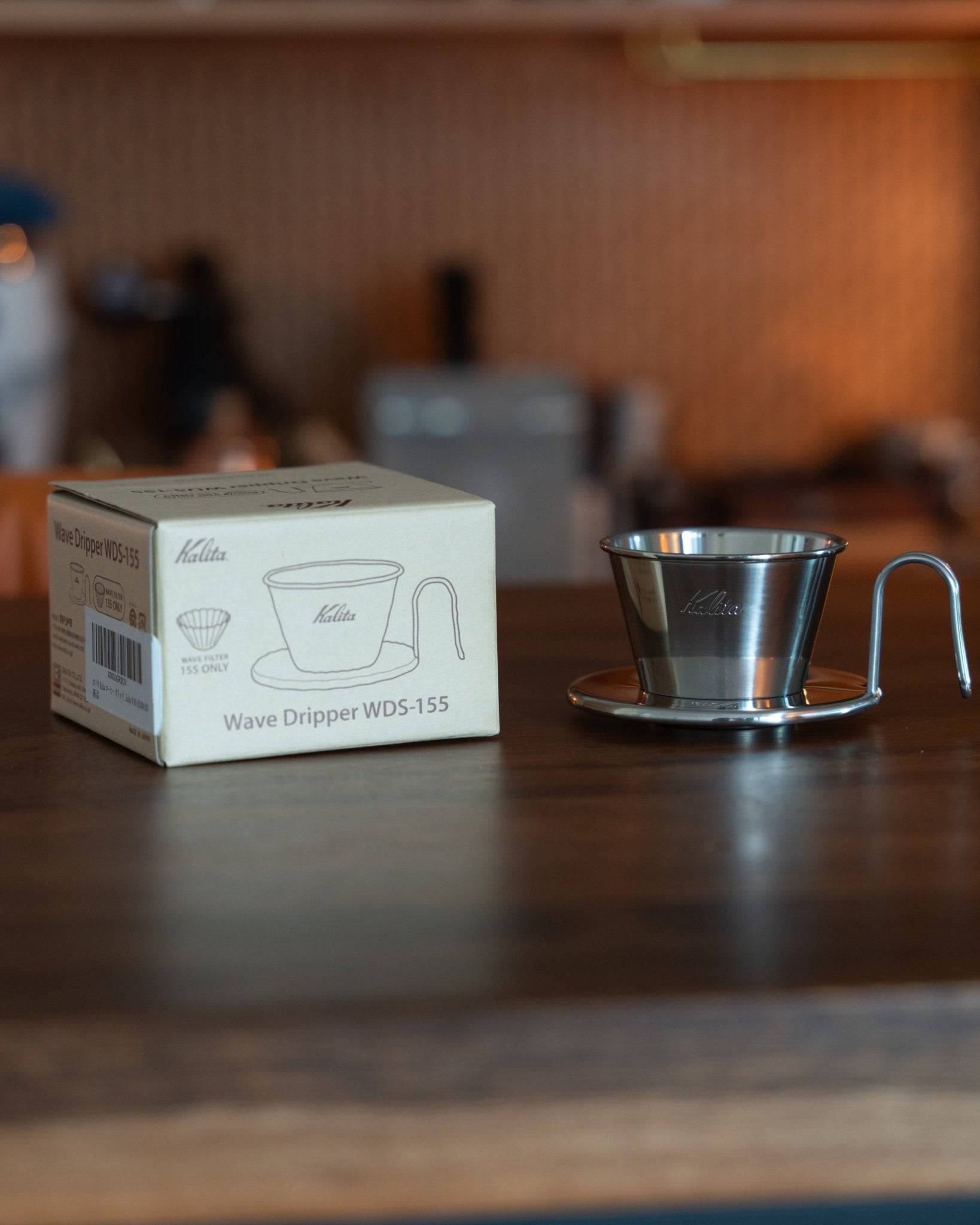 Kalita & TSUBAME WDS-155 stainless steel coffee dripper with product box, displayed on wooden table in coffee shop setting, showcasing elegant design and Japanese craftsmanship for premium pour-over brewing