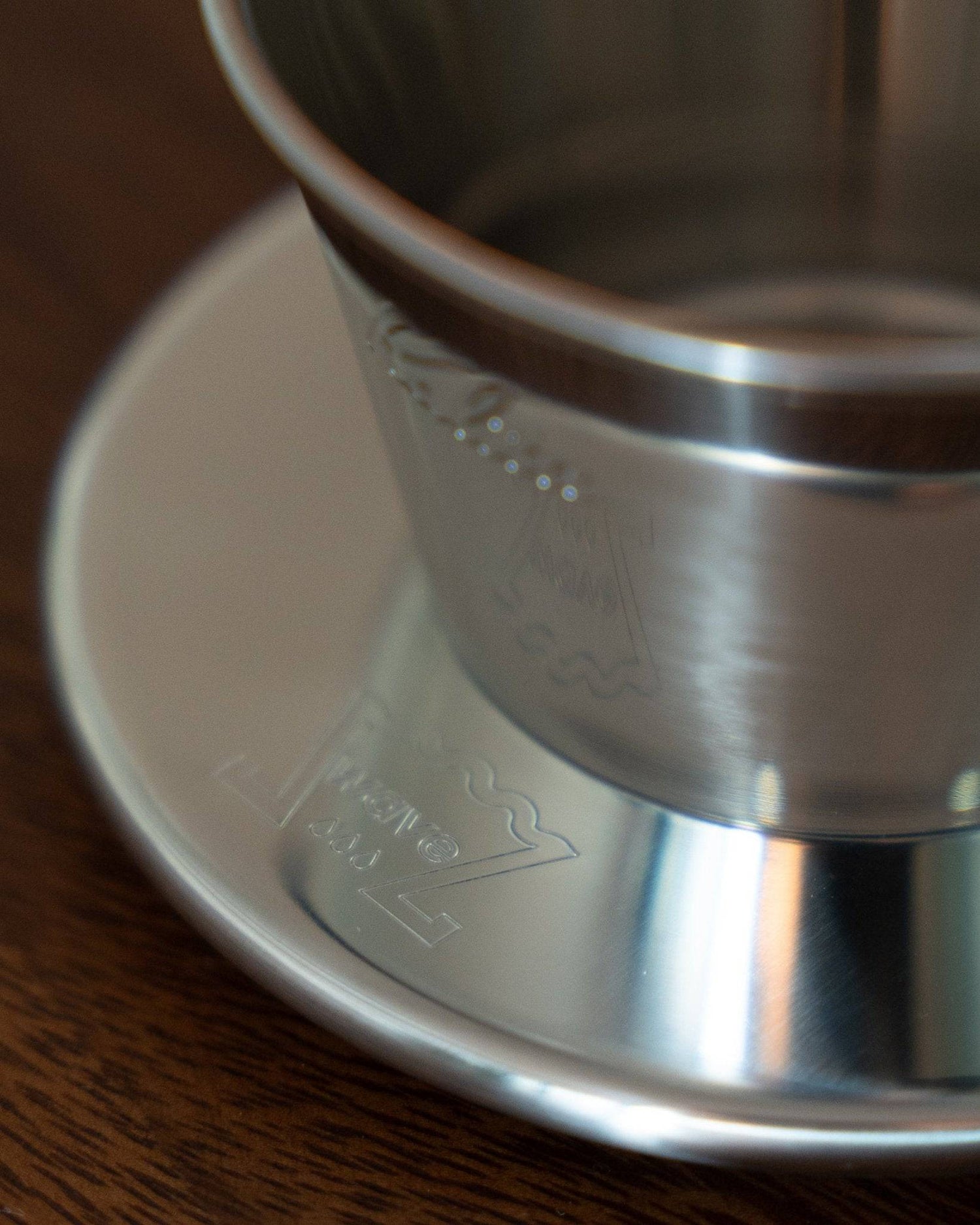 Kalita & TSUBAME WDS-155 stainless steel coffee dripper close-up, showing sleek design and engraved logo on brushed metal surface, placed on a white saucer atop wooden table