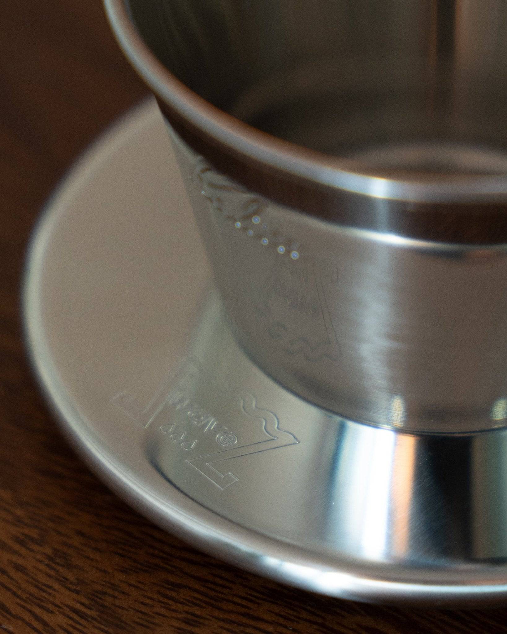 Kalita & TSUBAME WDS-155 stainless steel coffee dripper close-up, showing sleek design and engraved logo on brushed metal surface, placed on a white saucer atop wooden table