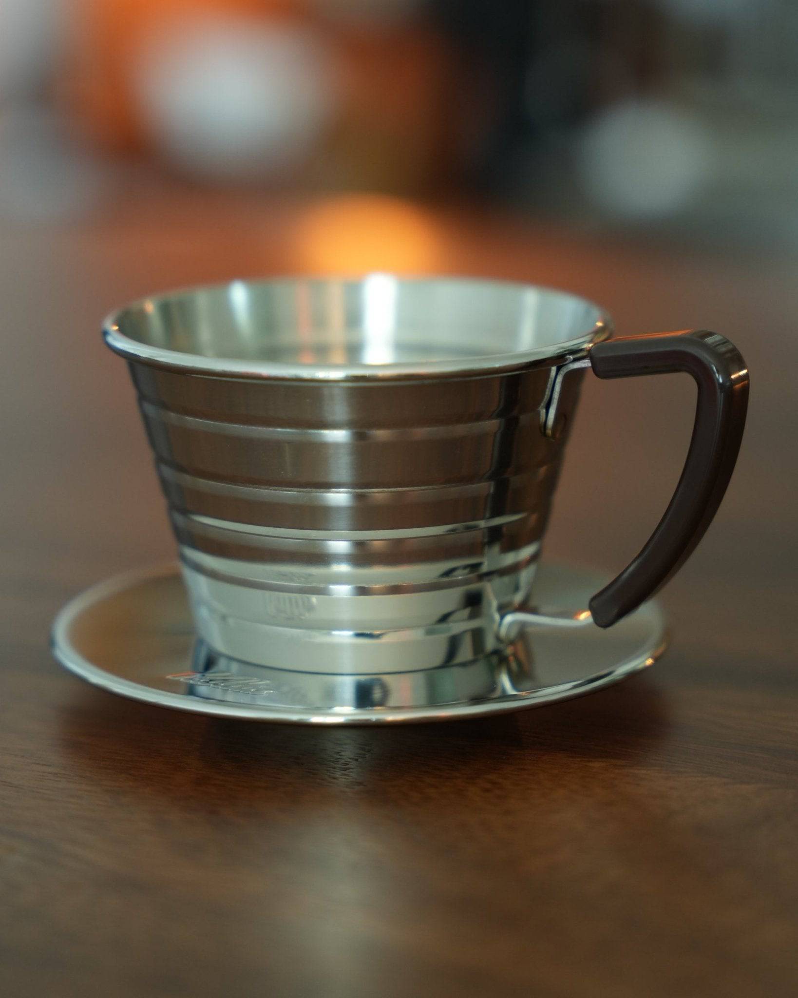 Kalita Wave 155 S stainless steel pour-over coffee dripper with unique wave pattern design, featuring a sleek handle and saucer, positioned on a wooden surface with soft background blur