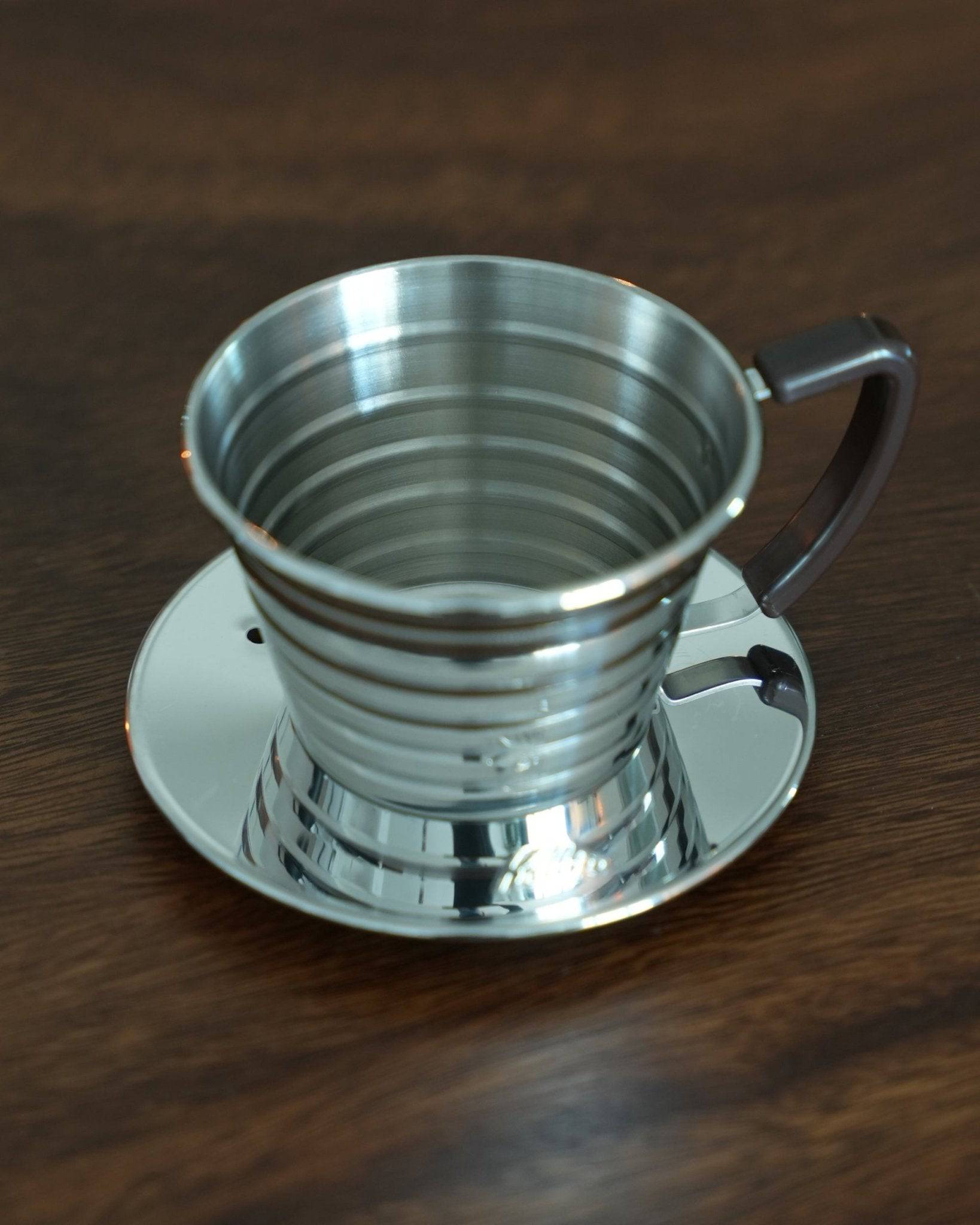 Kalita Wave 155 S stainless steel pour-over coffee dripper with distinctive wave pattern design, featuring a handle and saucer, placed on a wooden surface, ideal for brewing premium hand-drip coffee