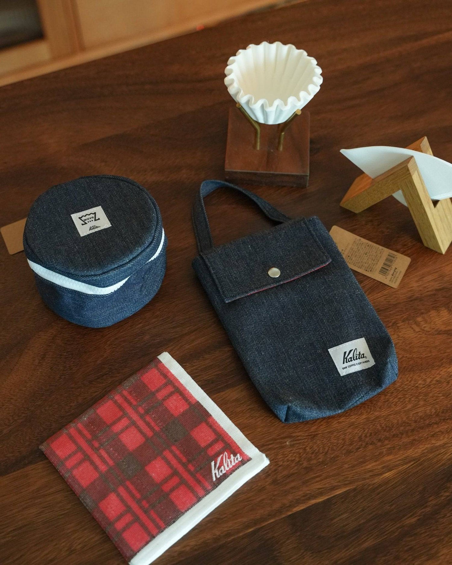 Kalita Wave coffee accessories on wooden table including denim filter storage bag, plaid pattern filter holder, circular container, and coffee dripper with stand