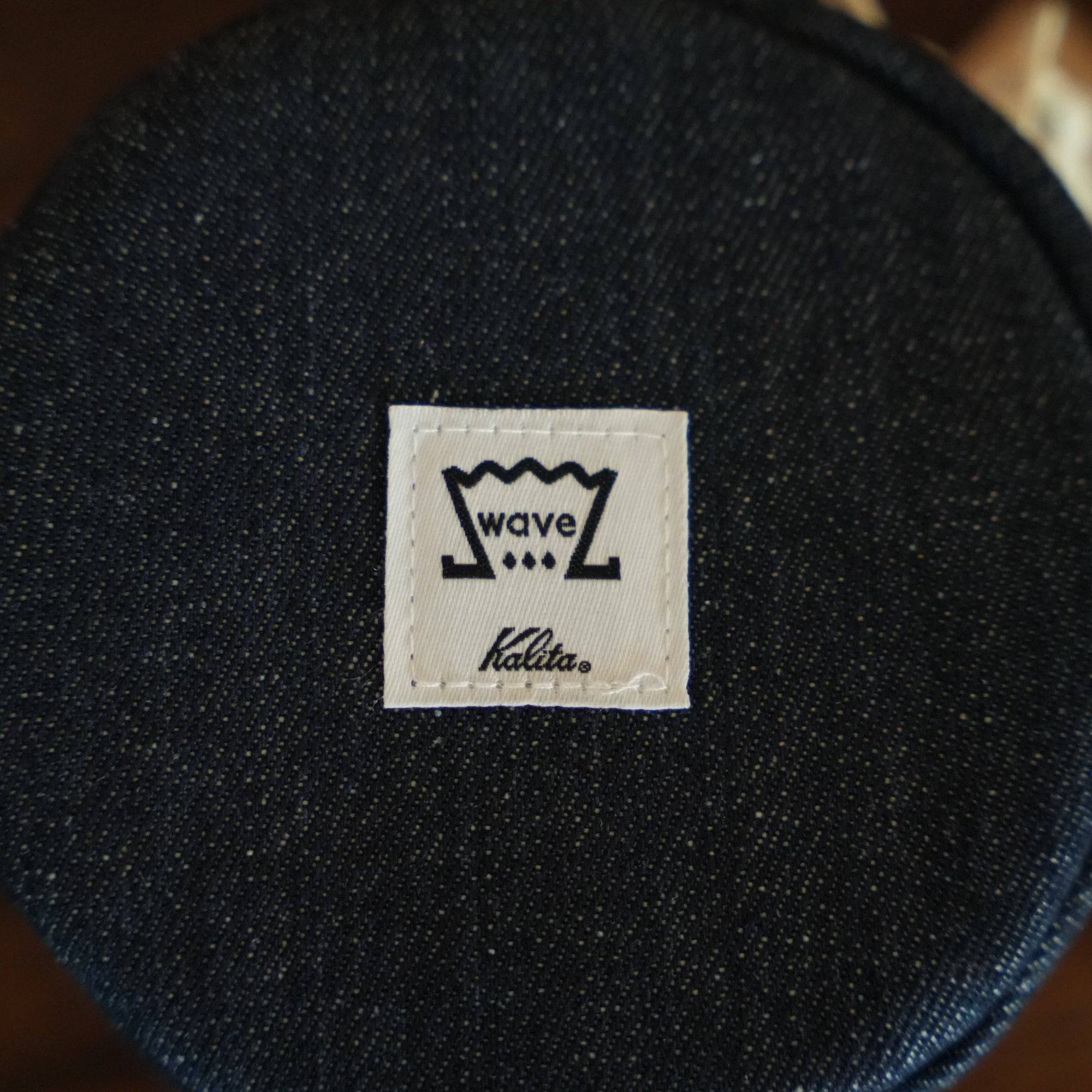 Close-up of Kalita Wave logo on dark denim fabric, showing stylized wave design and brand name on a square white patch stitched to the material, likely part of a coffee filter storage bag.