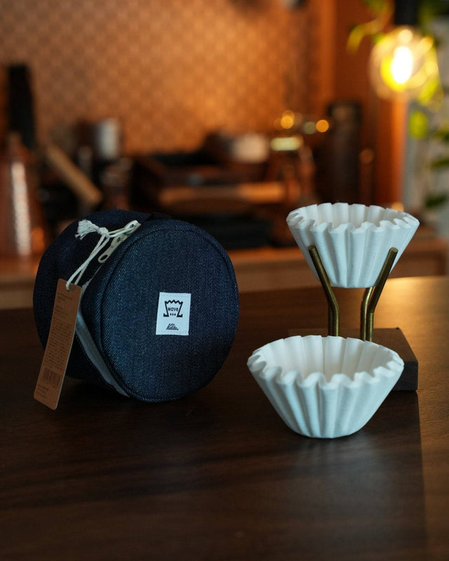 Kalita Wave coffee filter storage bag with white coffee filters on wooden table, warm cafe background