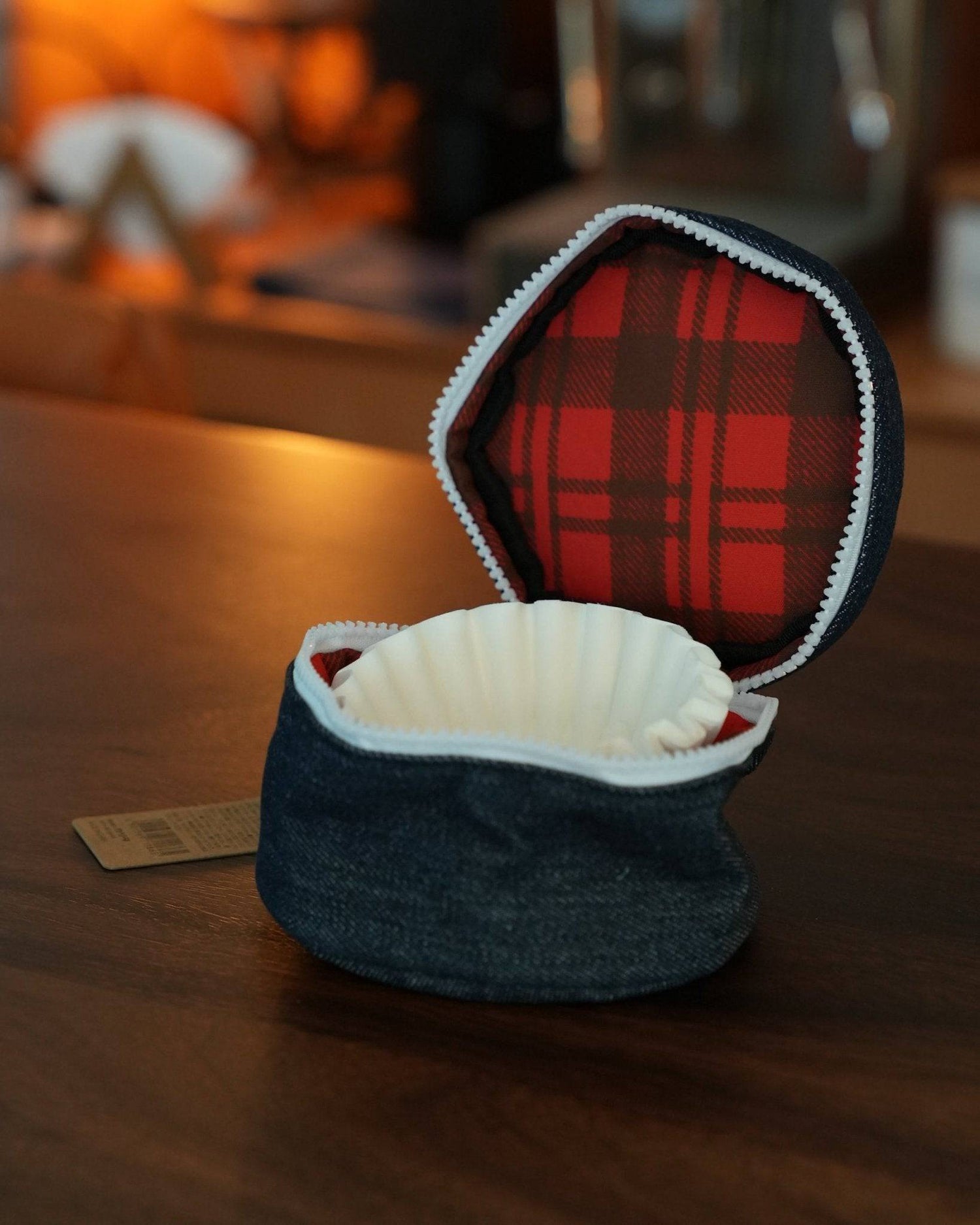 Kalita Wave coffee filter storage bag open on wooden table, showing red plaid interior lining and white filters inside. Denim exterior with zipper closure visible.