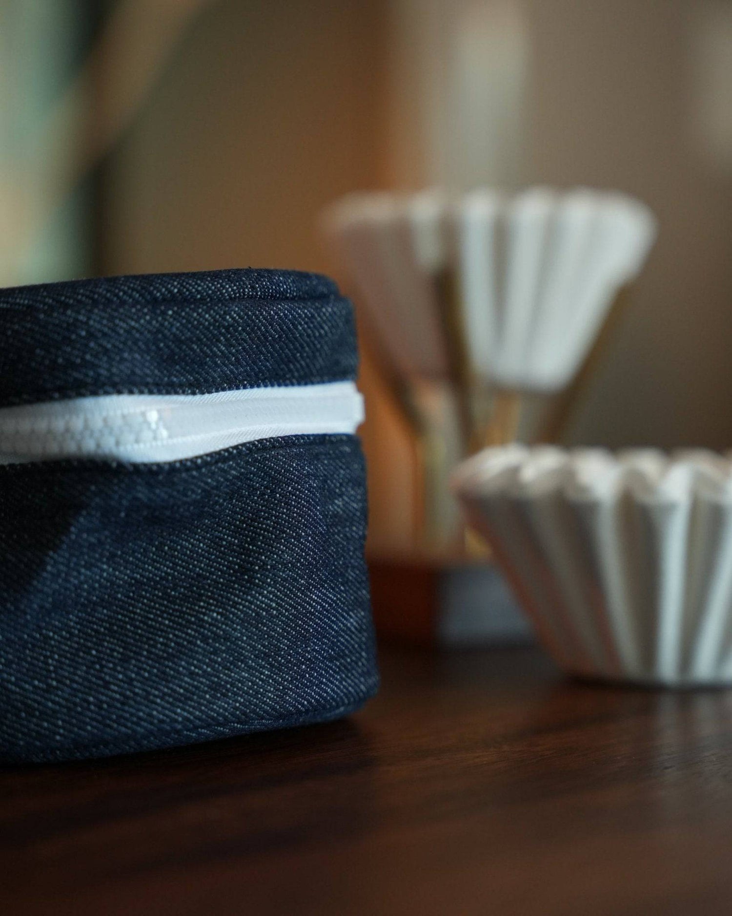 Kalita Wave coffee filter storage bag in denim blue with white zipper, displayed next to white paper coffee filters on wooden surface, showcasing stylish and practical design for coffee enthusiasts