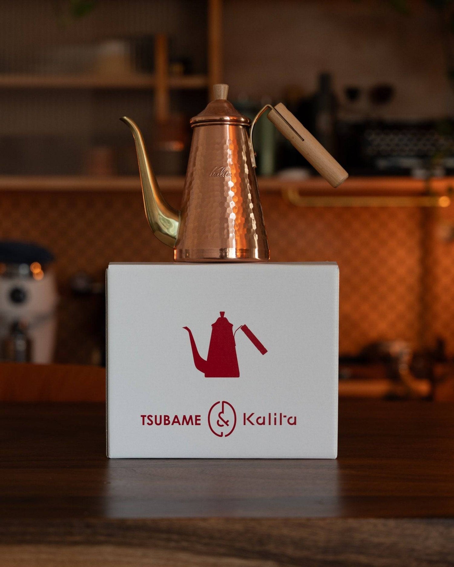 Copper Kalita x Tsubame kettle with long spout and wooden handle displayed on white box with red logo, showcasing Japanese craftsmanship and premium coffee brewing equipment in warm, rustic kitchen setting