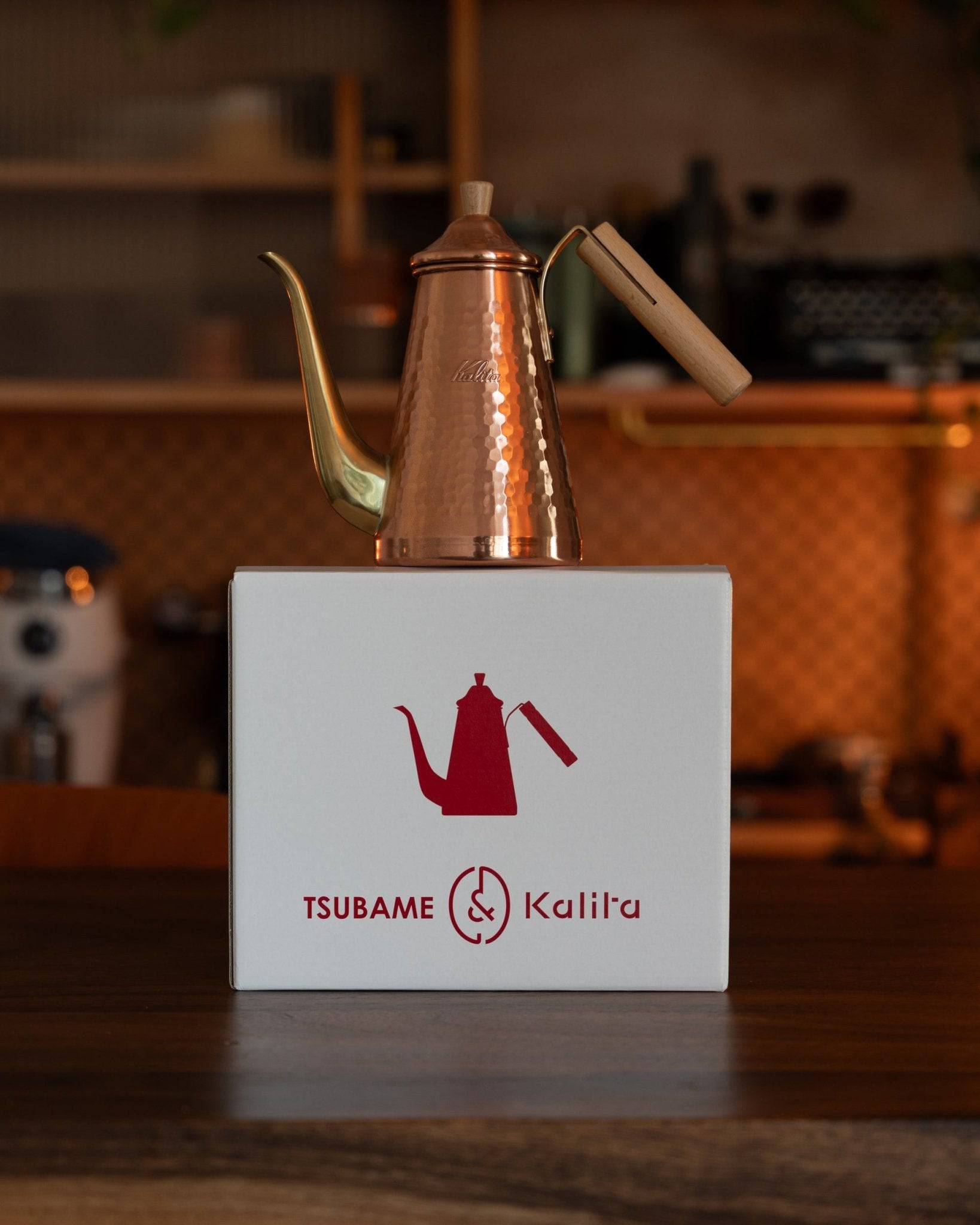 Copper Kalita x Tsubame kettle with long spout and wooden handle displayed on white box with red logo, showcasing Japanese craftsmanship and premium coffee brewing equipment in warm, rustic kitchen setting