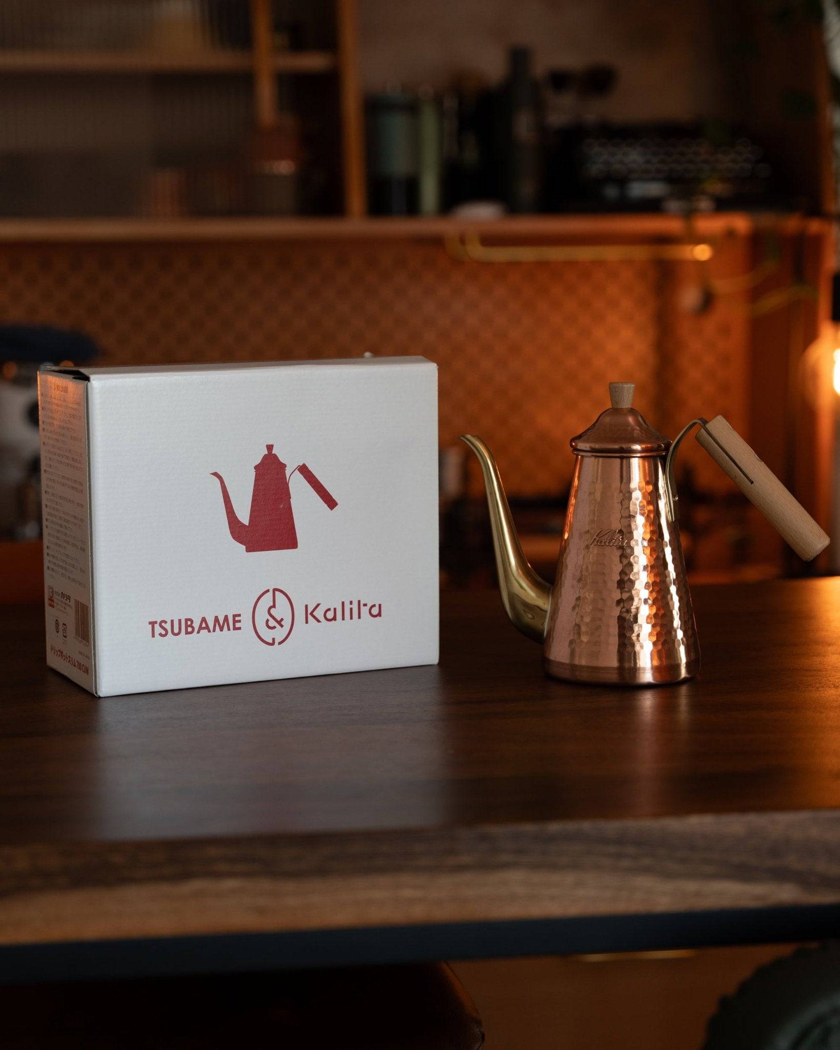 Kalita x Tsubame copper pour-over kettle with gooseneck spout and wooden handle, displayed next to its branded packaging box on a dark wooden surface, warm lighting highlights the kettle&