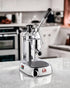 La Pavoni Europiccola Lusso lever espresso machine on kitchen counter, featuring chrome body, black handle, and drip tray. Professional-grade manual coffee maker for home baristas.