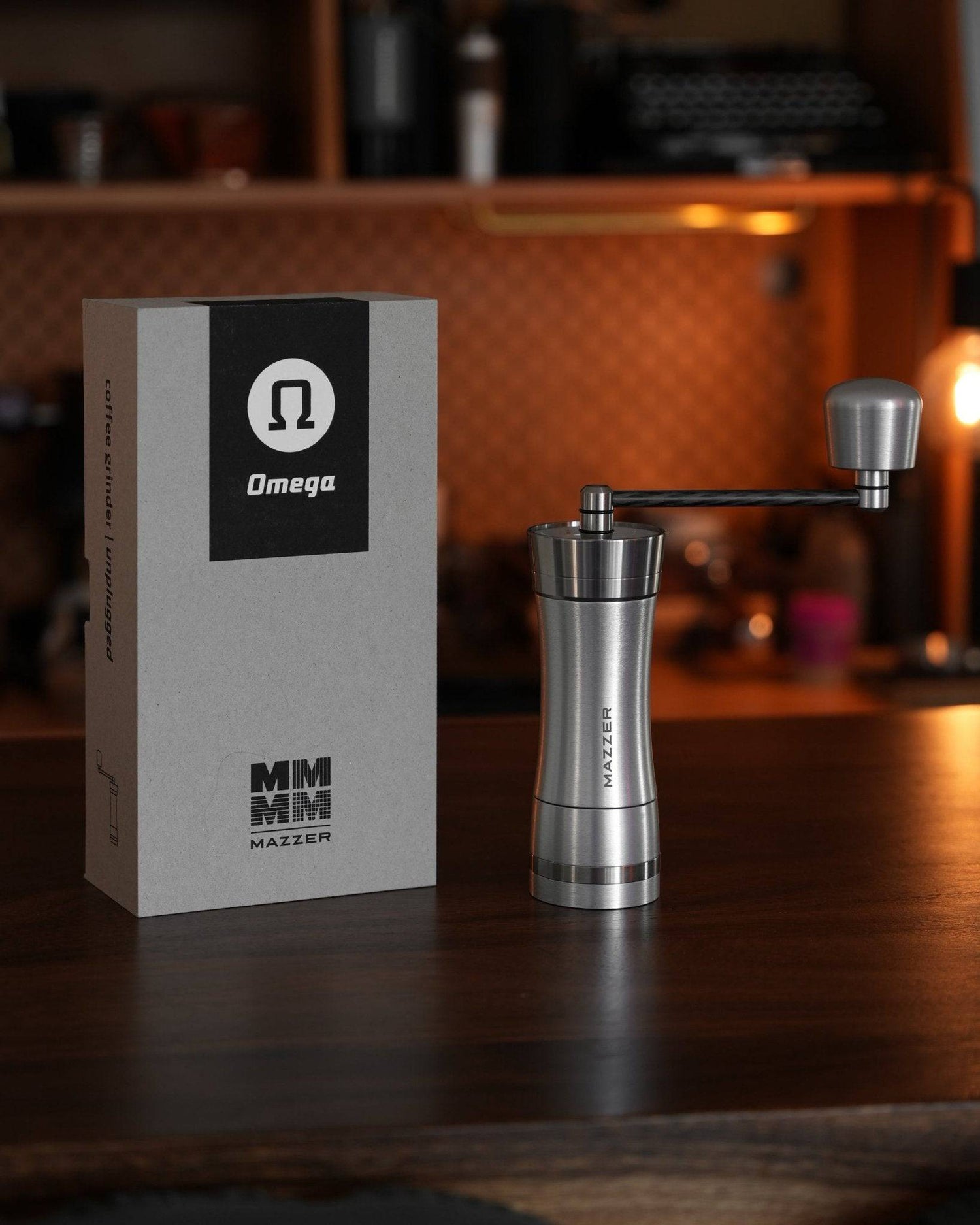 Mazzer Omega Fast manual coffee grinder with its packaging box, displayed on wooden counter in warm, cozy coffee shop setting. Stainless steel grinder body with ergonomic handle and adjustment knob visible.