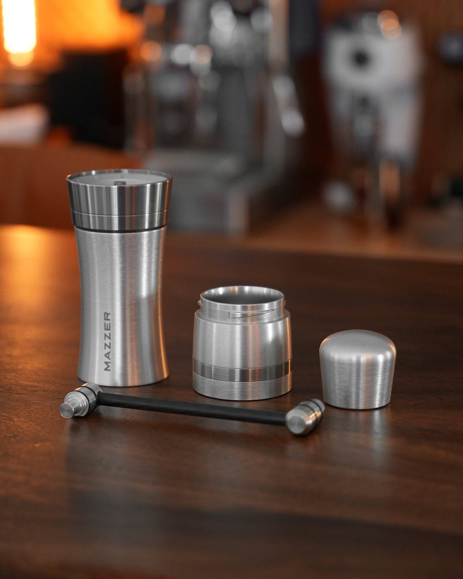 Mazzer Omega Soft manual coffee grinder disassembled on wooden counter, showing stainless steel body, grind adjustment ring, and hand crank, with warm lighting and coffee equipment in background