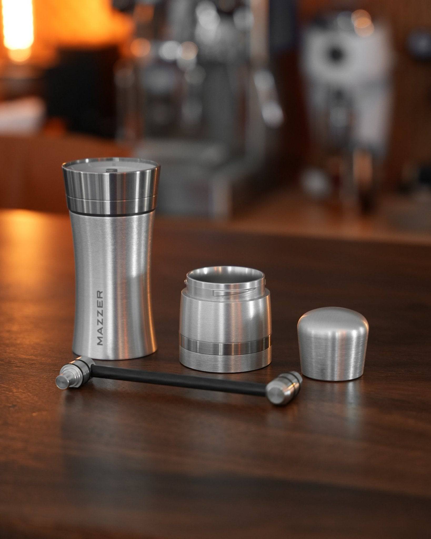 Mazzer Omega Soft manual coffee grinder disassembled on wooden counter, showing stainless steel body, grind adjustment ring, and hand crank, with warm lighting and coffee equipment in background