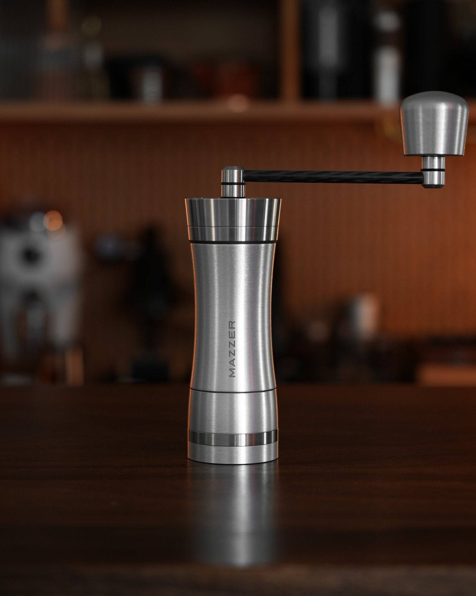 Mazzer Omega Soft manual coffee grinder on wooden table, sleek stainless steel design with carbon fiber handle, conical burrs for precision grinding, compact and portable for home or travel use
