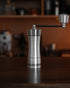 Mazzer Omega Soft manual coffee grinder on wooden table, sleek stainless steel design with carbon fiber handle, conical burrs for precision grinding, compact and portable for home or travel use