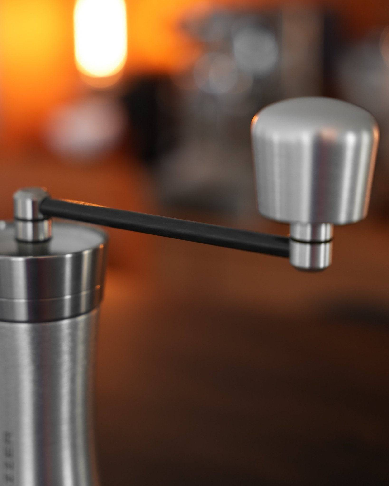 Mazzer Omega Soft manual coffee grinder with sleek stainless steel body, carbon fiber handle, and adjustable grind settings, displayed on warm wooden surface with soft orange background lighting