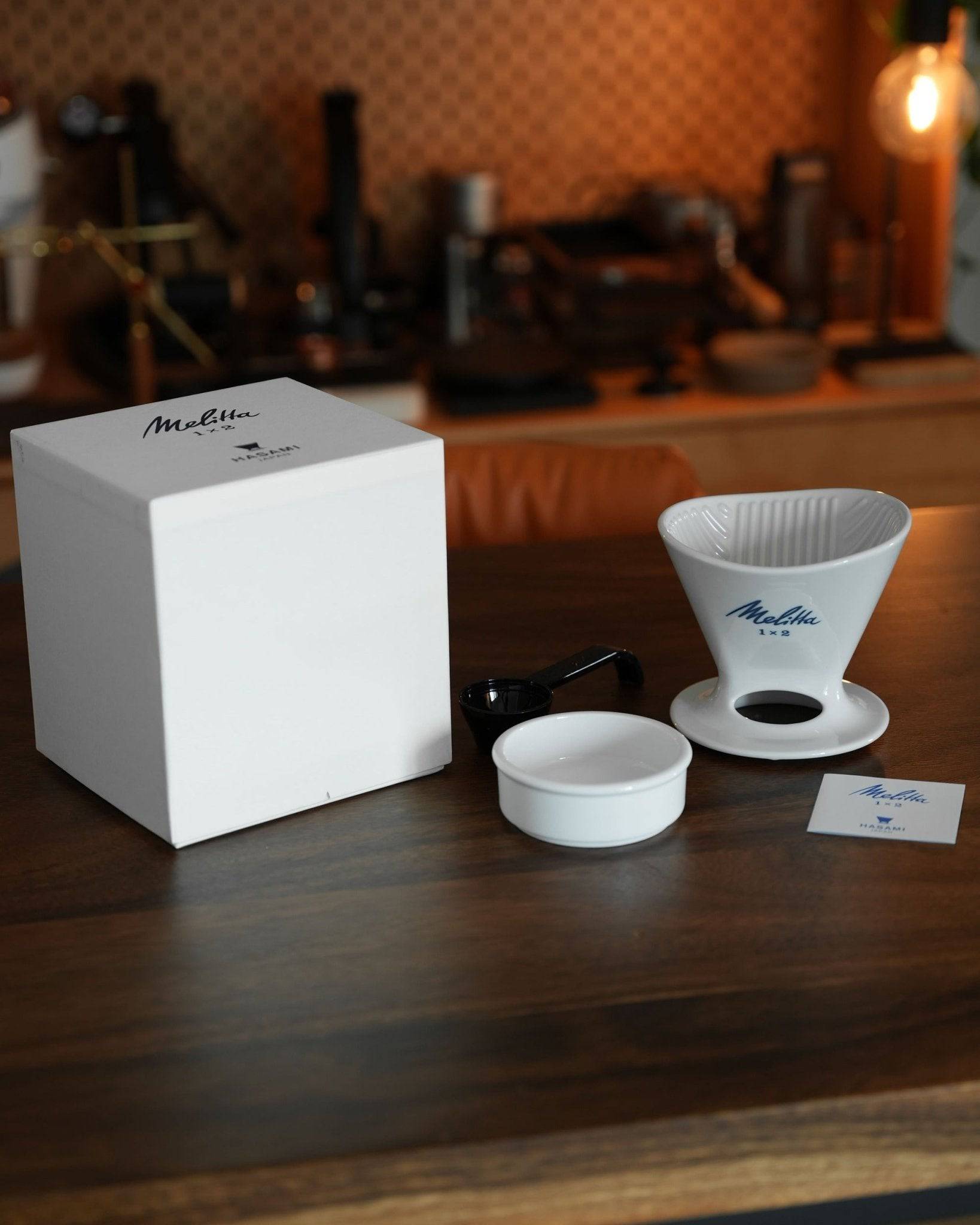 Melitta and HASAMI ceramic pour-over coffee dripper set with white box, filter cone, and saucer on wooden table, coffee equipment in background