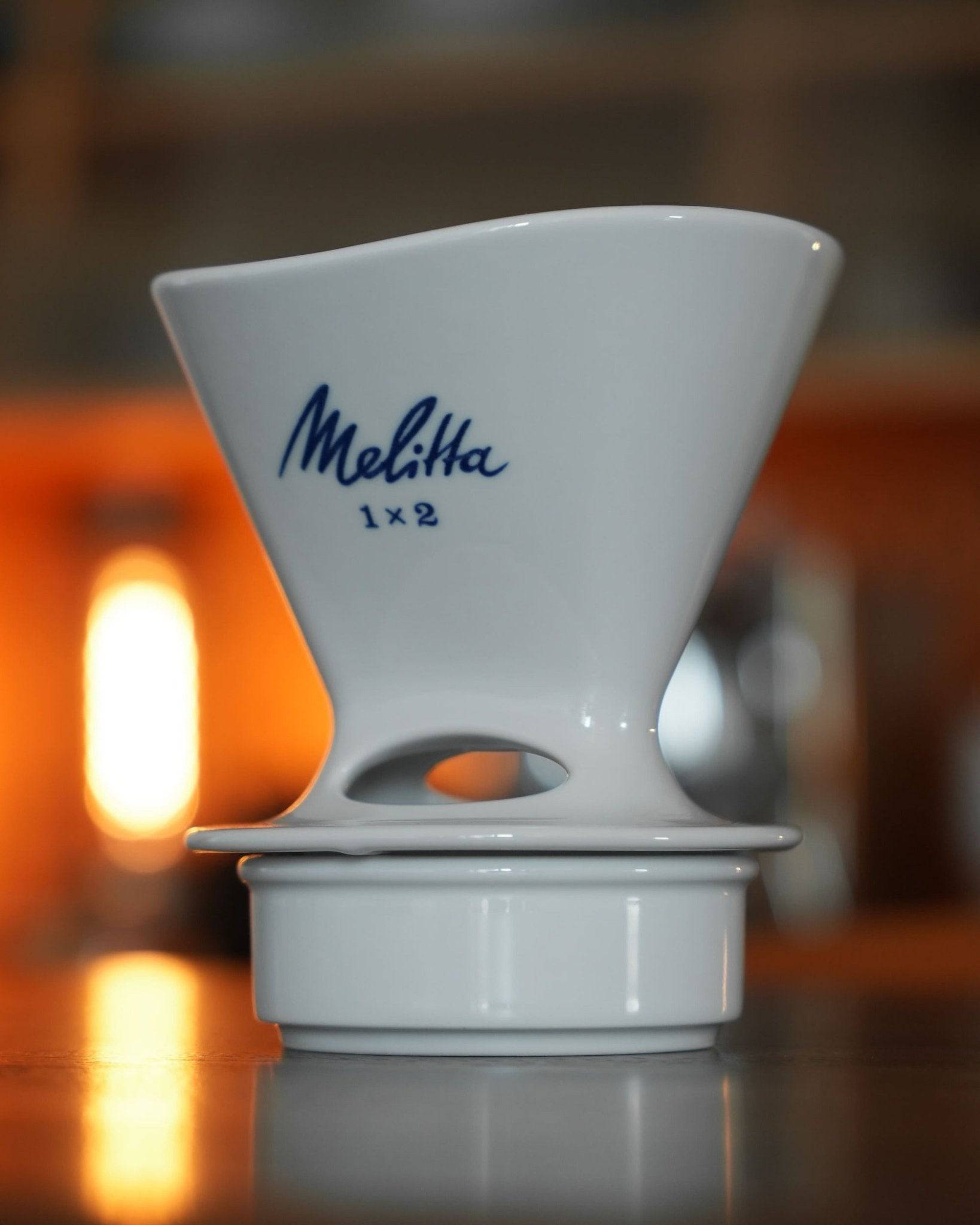 White ceramic Melitta 1x2 pour-over coffee dripper with blue logo, sitting on a matching white base, illuminated by warm light in background