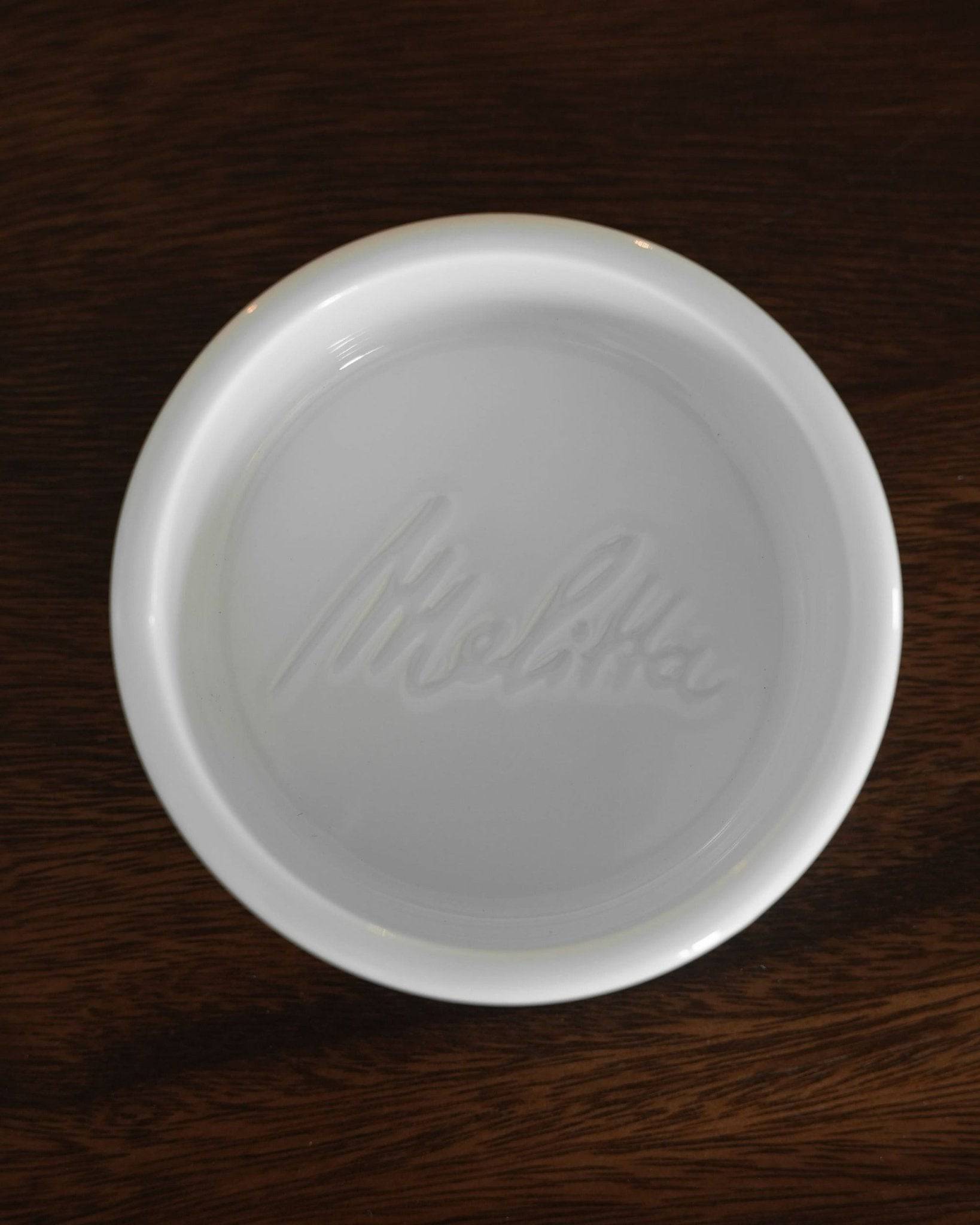 White ceramic dish with embossed Melitta logo, part of the limited edition Melitta & HASAMI ceramic pour-over set, displayed on dark wooden surface