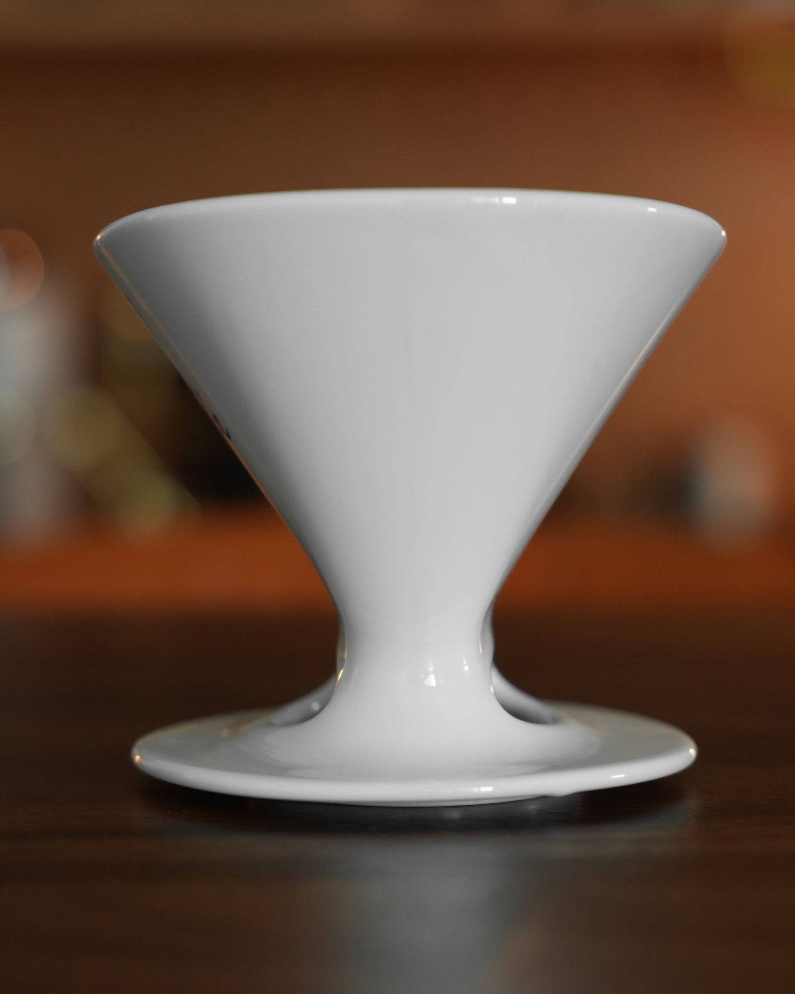 White ceramic pour-over coffee dripper with conical shape and ribbed interior, sitting on a saucer, showcasing Melitta and Hasami&