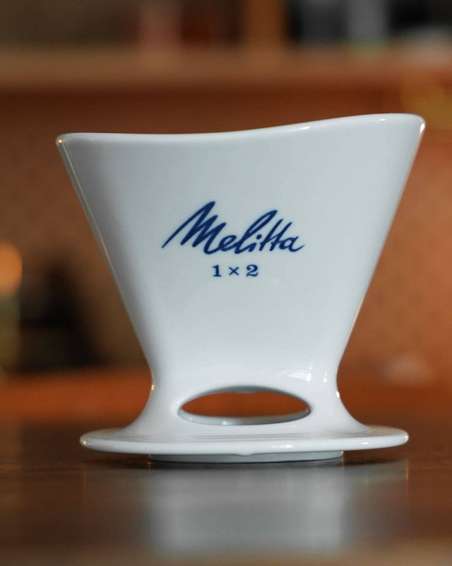 White ceramic Melitta 1x2 pour-over coffee dripper with blue logo, featuring asymmetrical design and single hole bottom, displayed on wooden surface