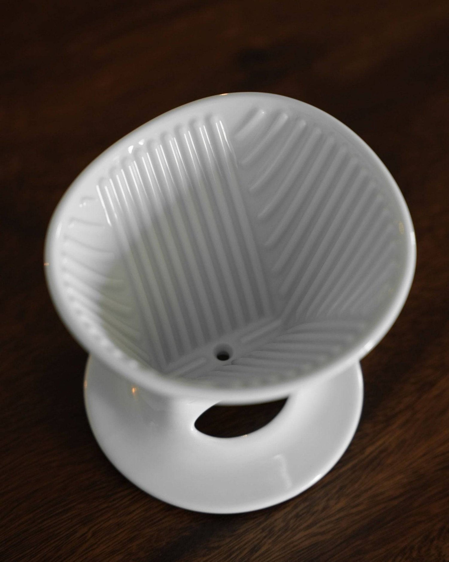 White ceramic pour-over coffee dripper with distinctive ribbed interior design, single hole at bottom, and attached saucer, showcasing Melitta and Hasami collaboration on dark wooden surface